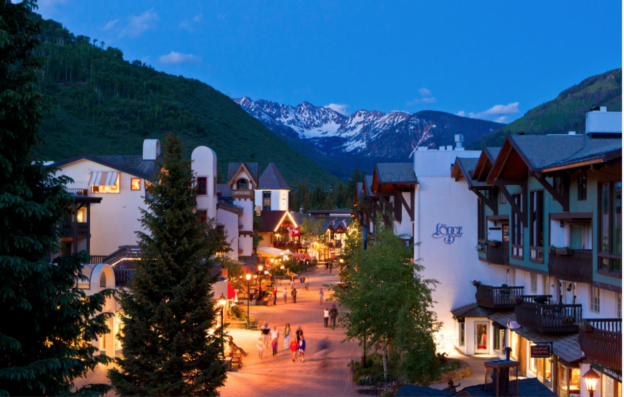 A lively mountain town during twilight with snow-capped peaks in the background, outdoor shops and cafes along a pedestrian street, and people walking and enjoying the evening.