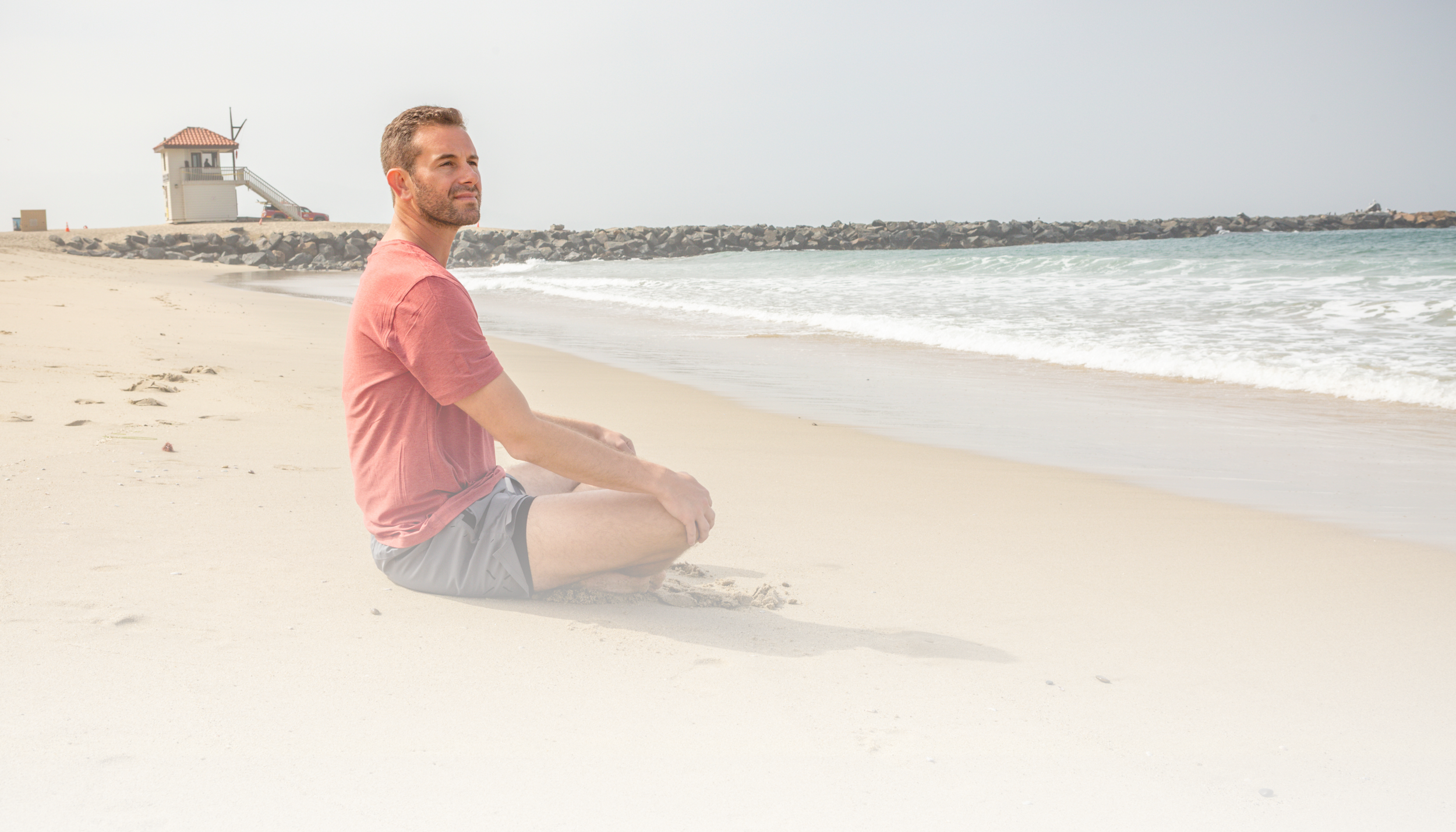 A man sitting cross-legged on a sandy beach, looking at the ocean with a jetty and small building in the background.