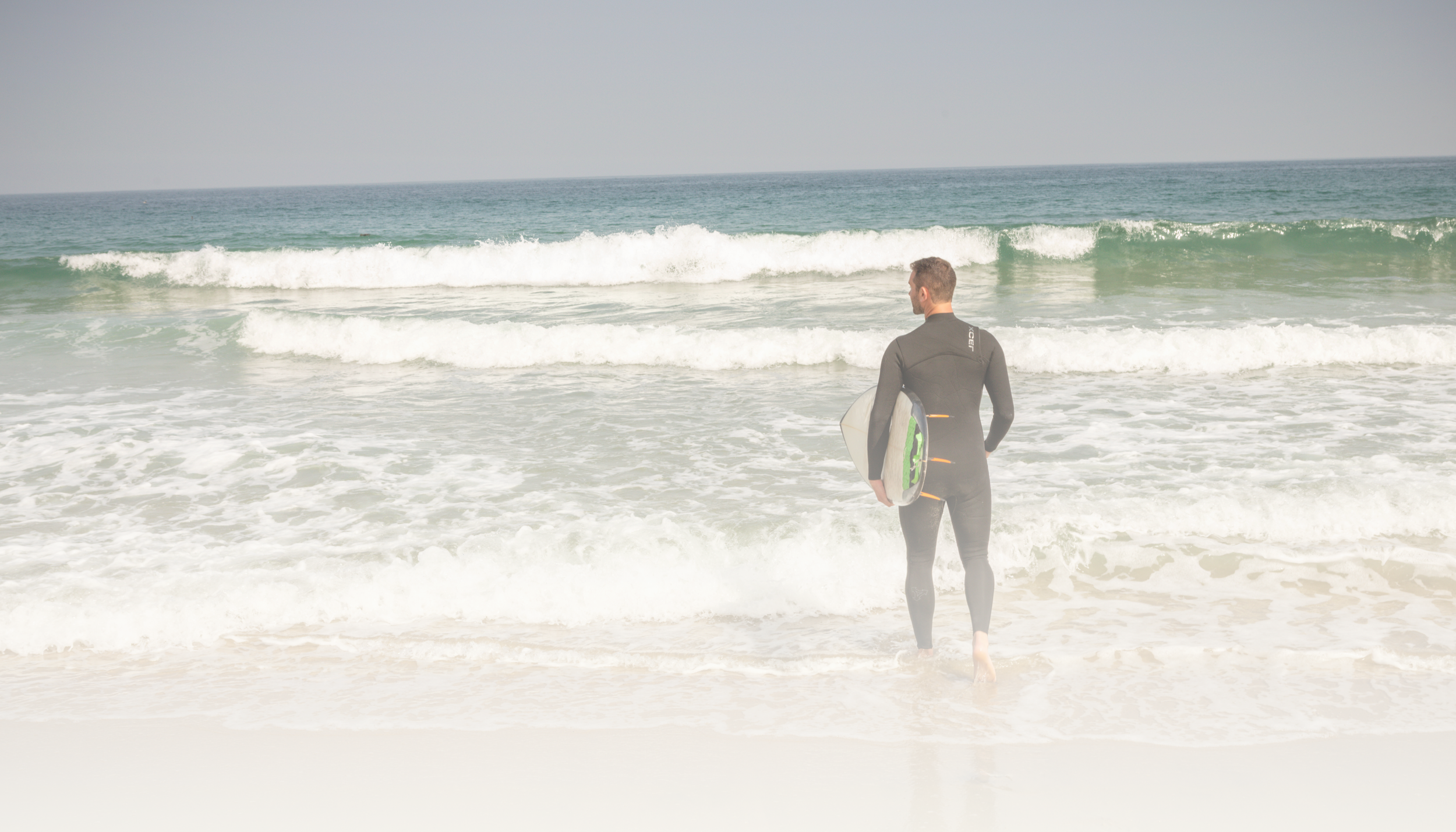 A man in a wetsuit holding a surfboard stands in the shallow ocean water and looks out towards the waves.
