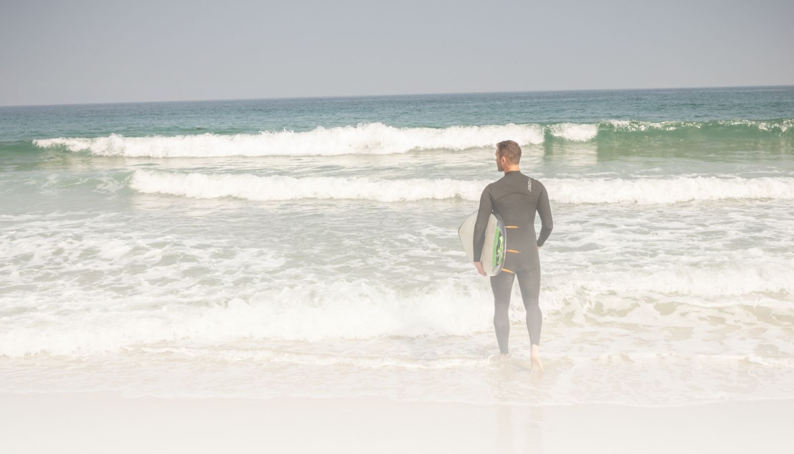A male surfer in a wetsuit walking into the ocean carrying a surfboard.