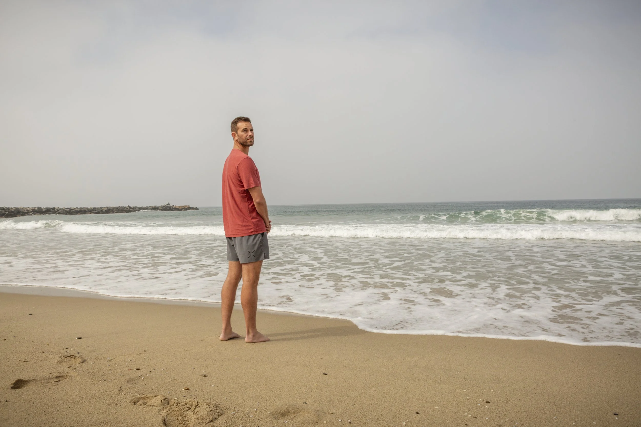 A man with short hair, wearing a coral t-shirt and gray shorts, standing barefoot on the sandy beach near the shoreline, looking towards the camera with the ocean and sky in the background.