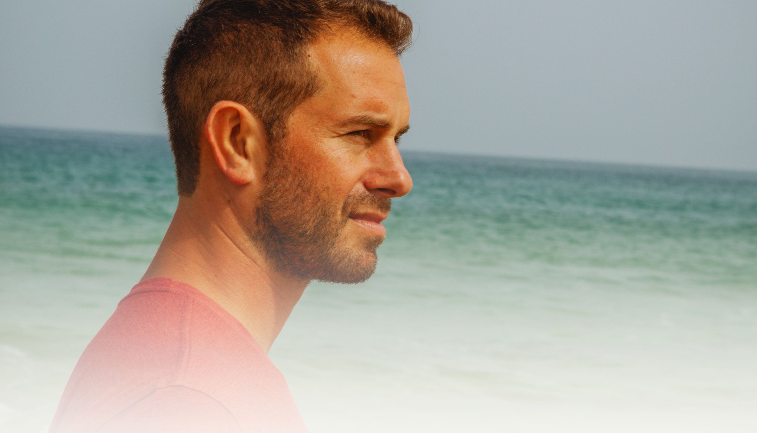 A man with short brown hair and a beard is standing on the beach, looking toward the ocean with a calm expression. The background features the sea and a pale sky.