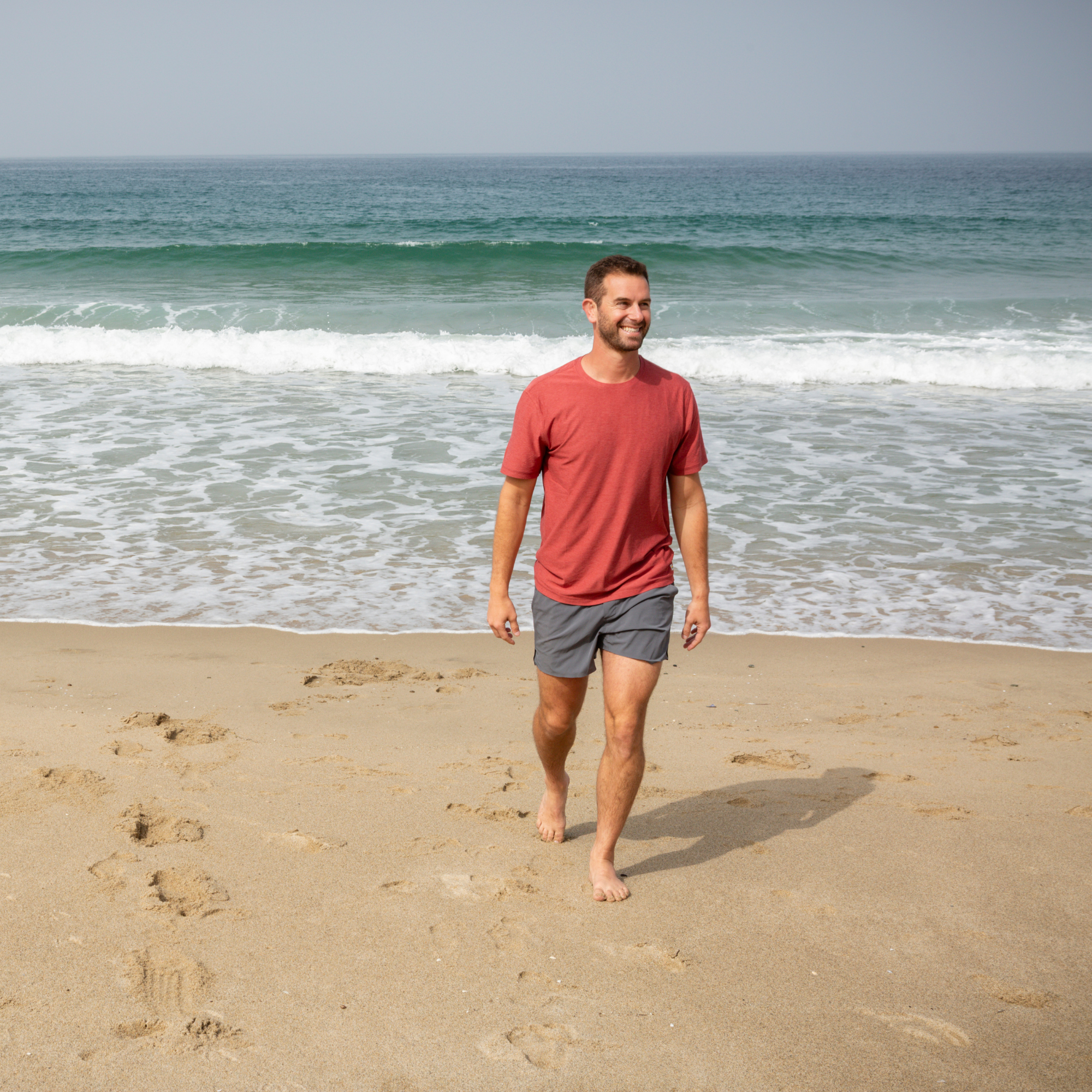 A man walking barefoot on the beach with the ocean in the background, smiling and wearing a red t-shirt and gray shorts.