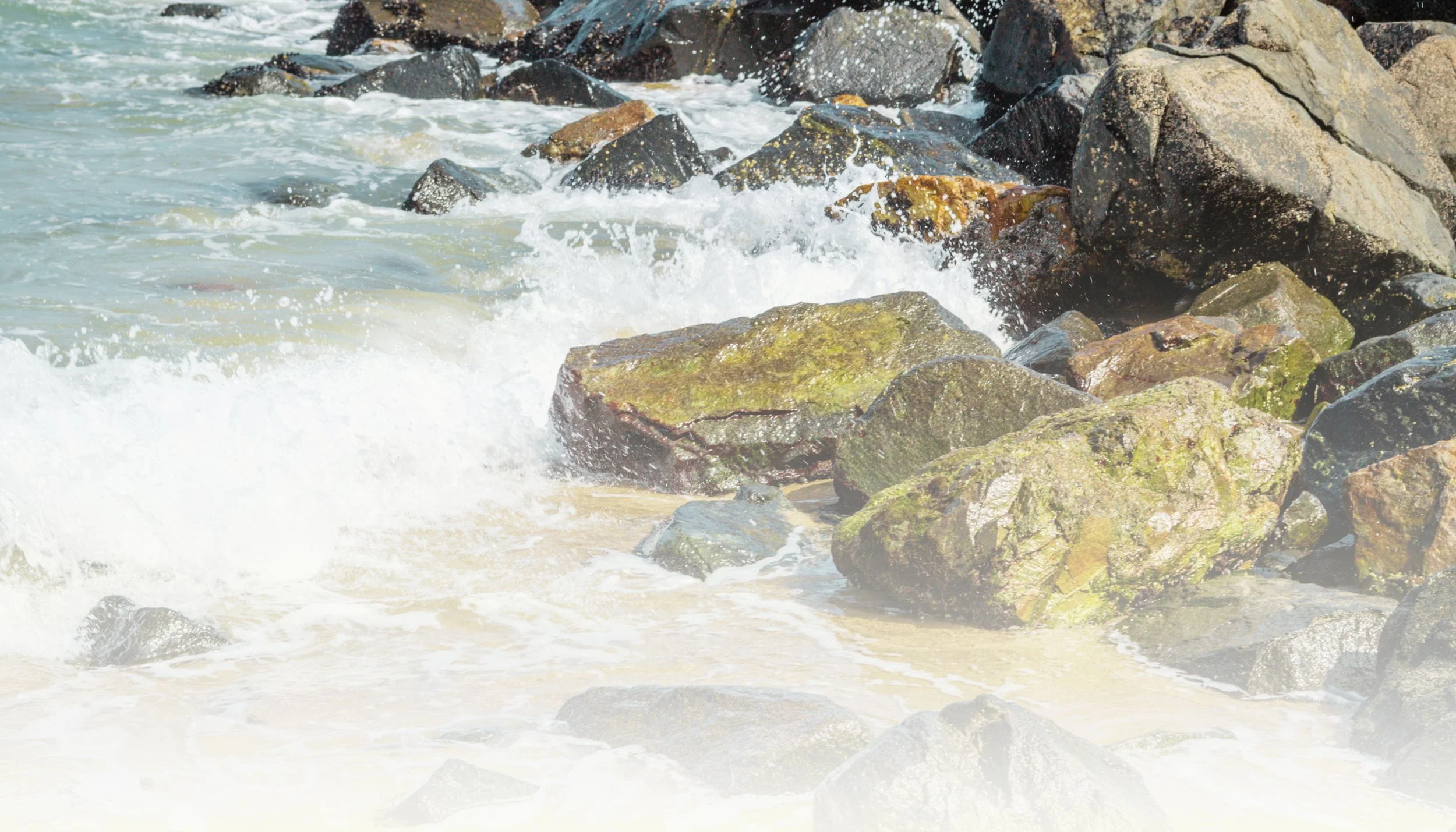 Waves crashing against rocks on a beach with foamy white water and some moss-covered stones.