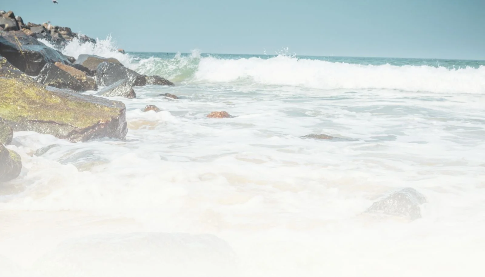 Ocean waves crashing against rocks on a sandy beach under a clear sky.