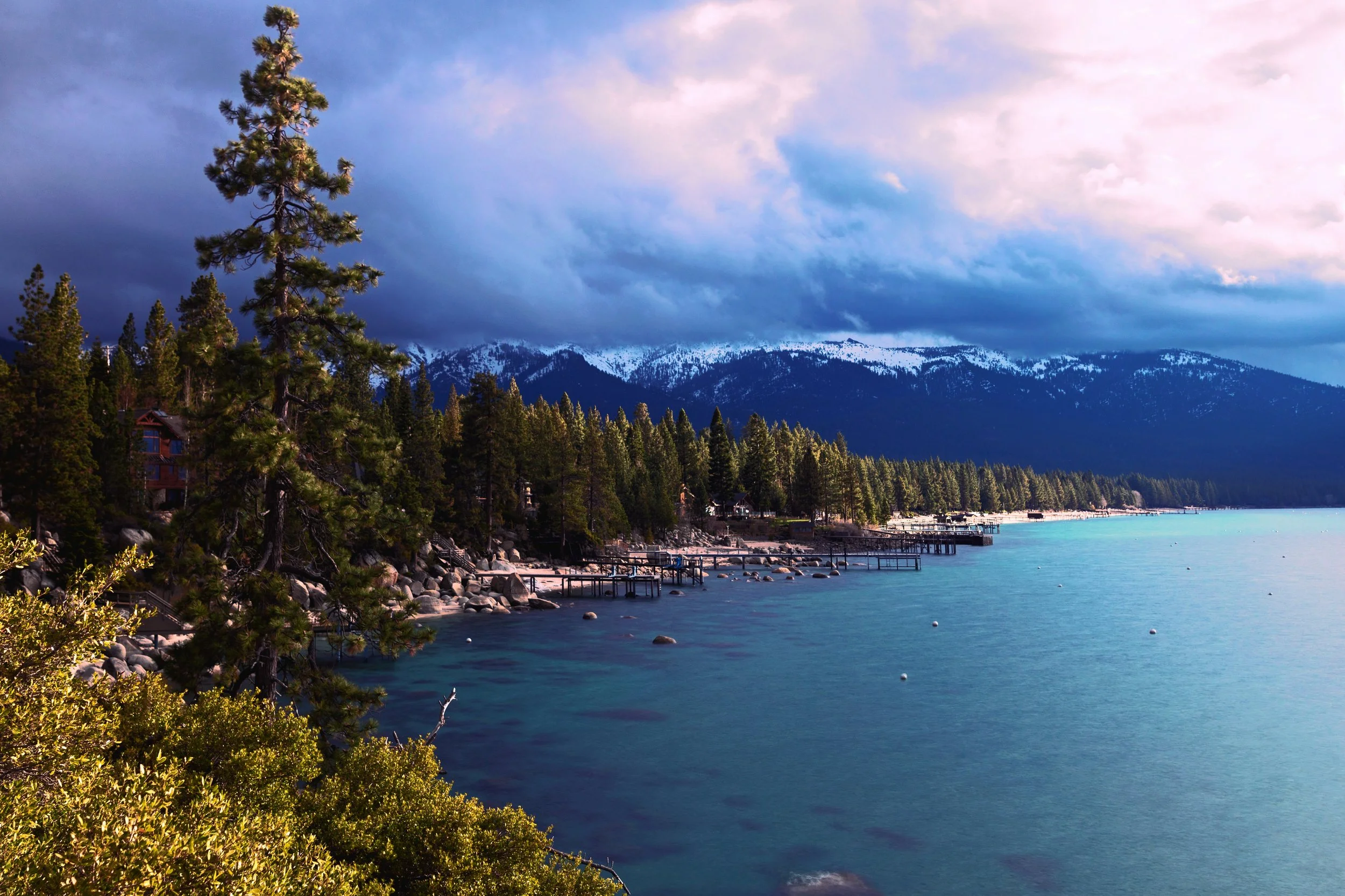 Lake with a dock and boats, surrounded by pine trees, with snow-capped mountains and a cloudy sky in the background.