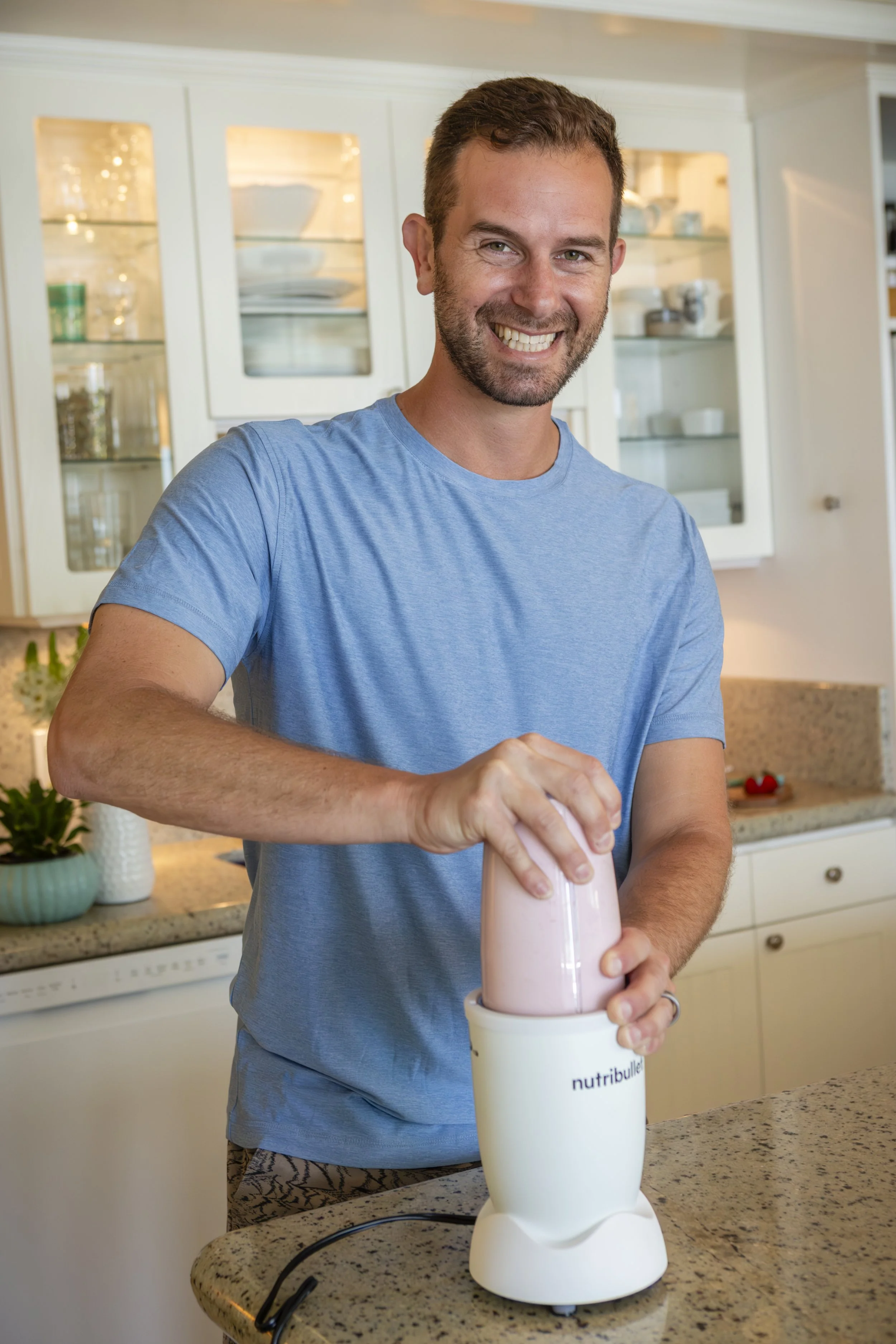 A man wearing a blue t-shirt is smiling as he prepares a NutriBullet blender with a light pink smoothie in a kitchen.