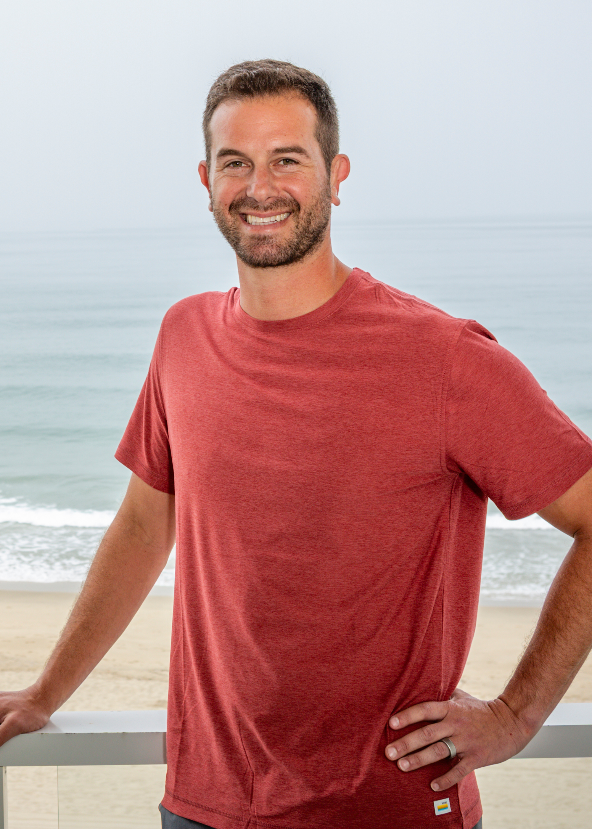 A man with short brown hair and a beard smiling, wearing a red T-shirt, standing near the beach with the ocean and sky in the background.