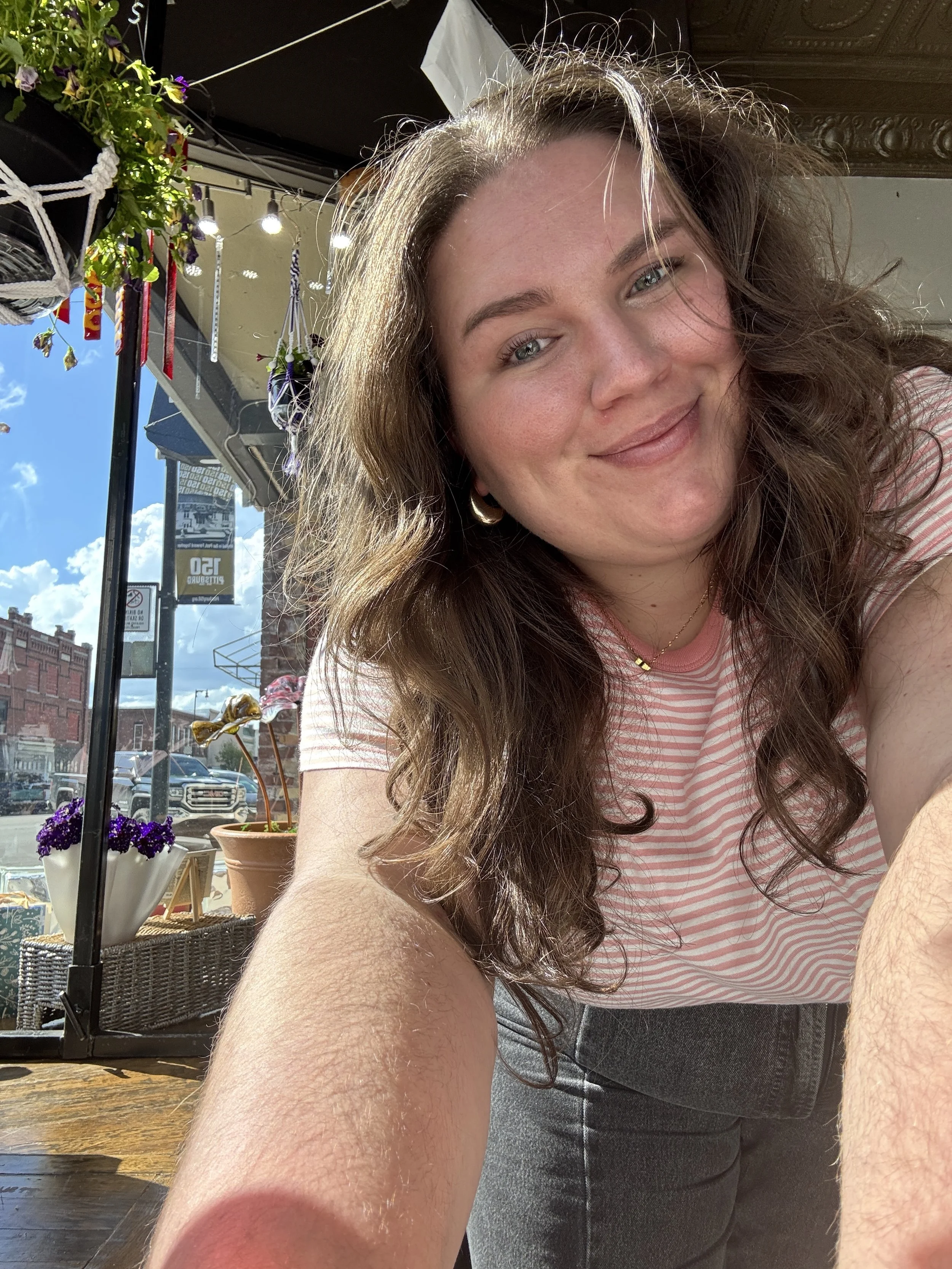 A woman with long wavy brown hair and wearing a pink striped t-shirt takes a selfie outdoors in sunlight, smiling with a city street scene in the background including potted plants, flowers, and storefronts.