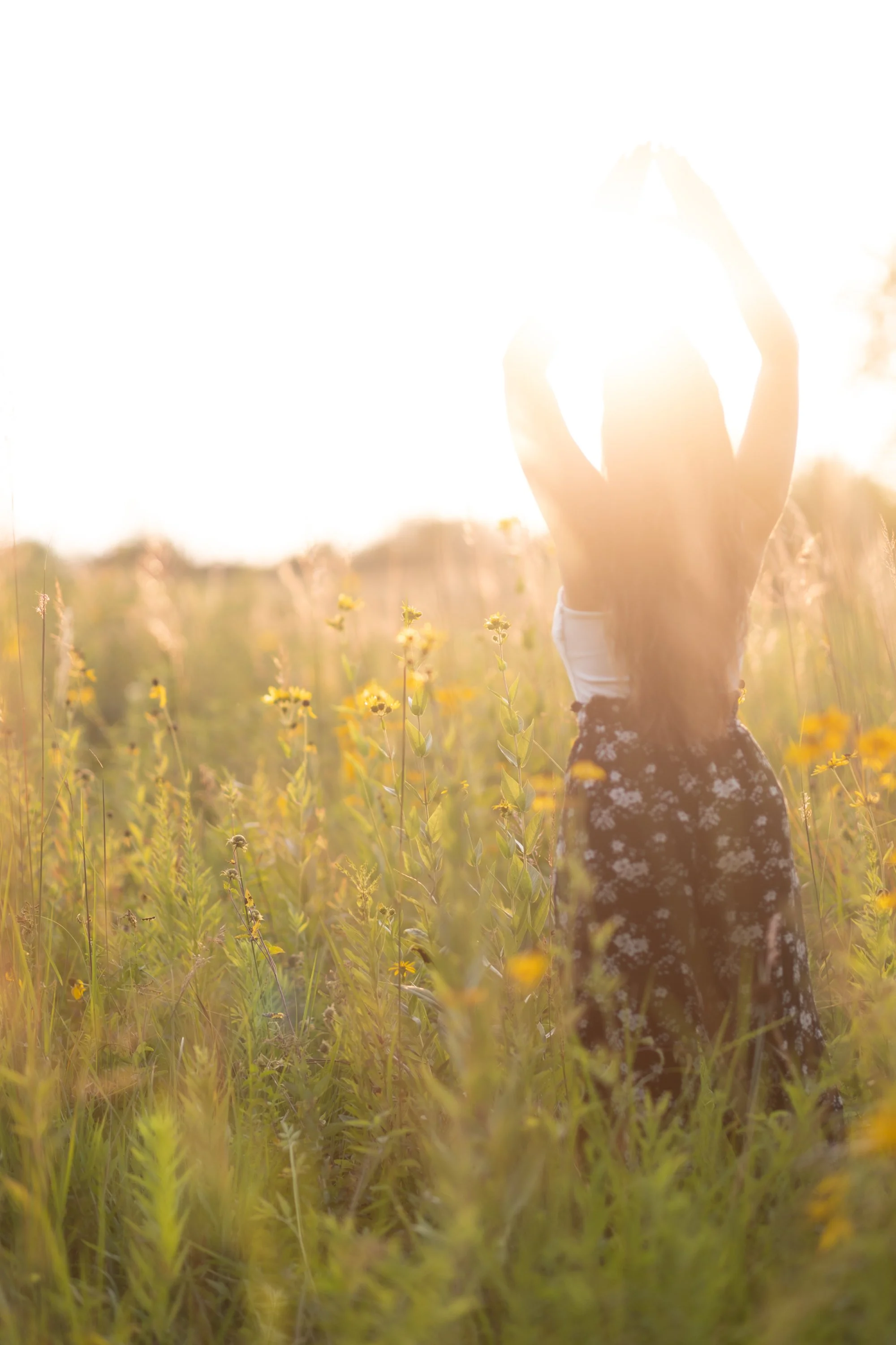 Person, Alicia, in a long floral dress standing in a field of yellow flowers at sunset, with arms raised and hands clasped above their head, back to the camera.
