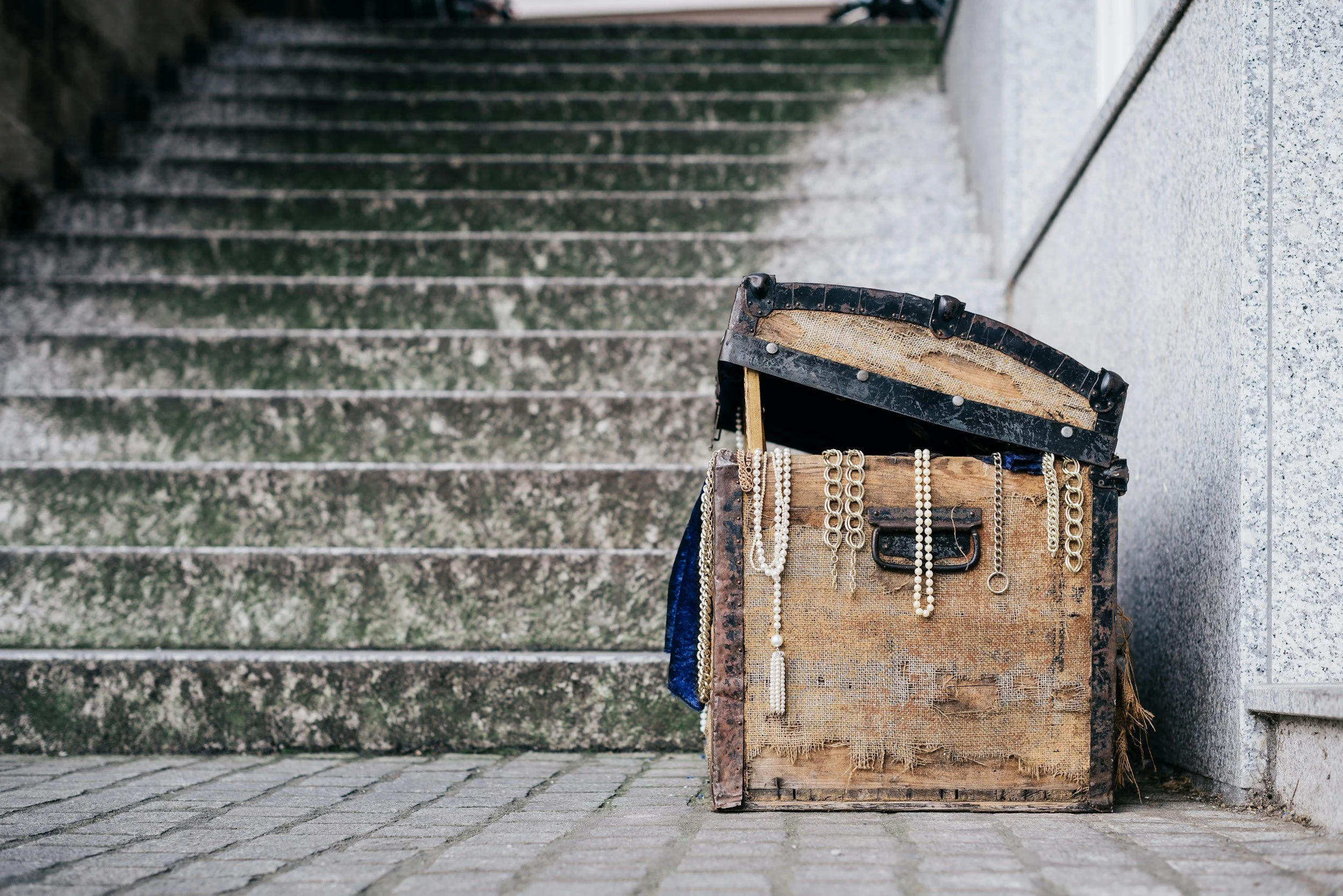 An open vintage suitcase filled with jewelry, placed on the ground beside stone stairs and a concrete wall.