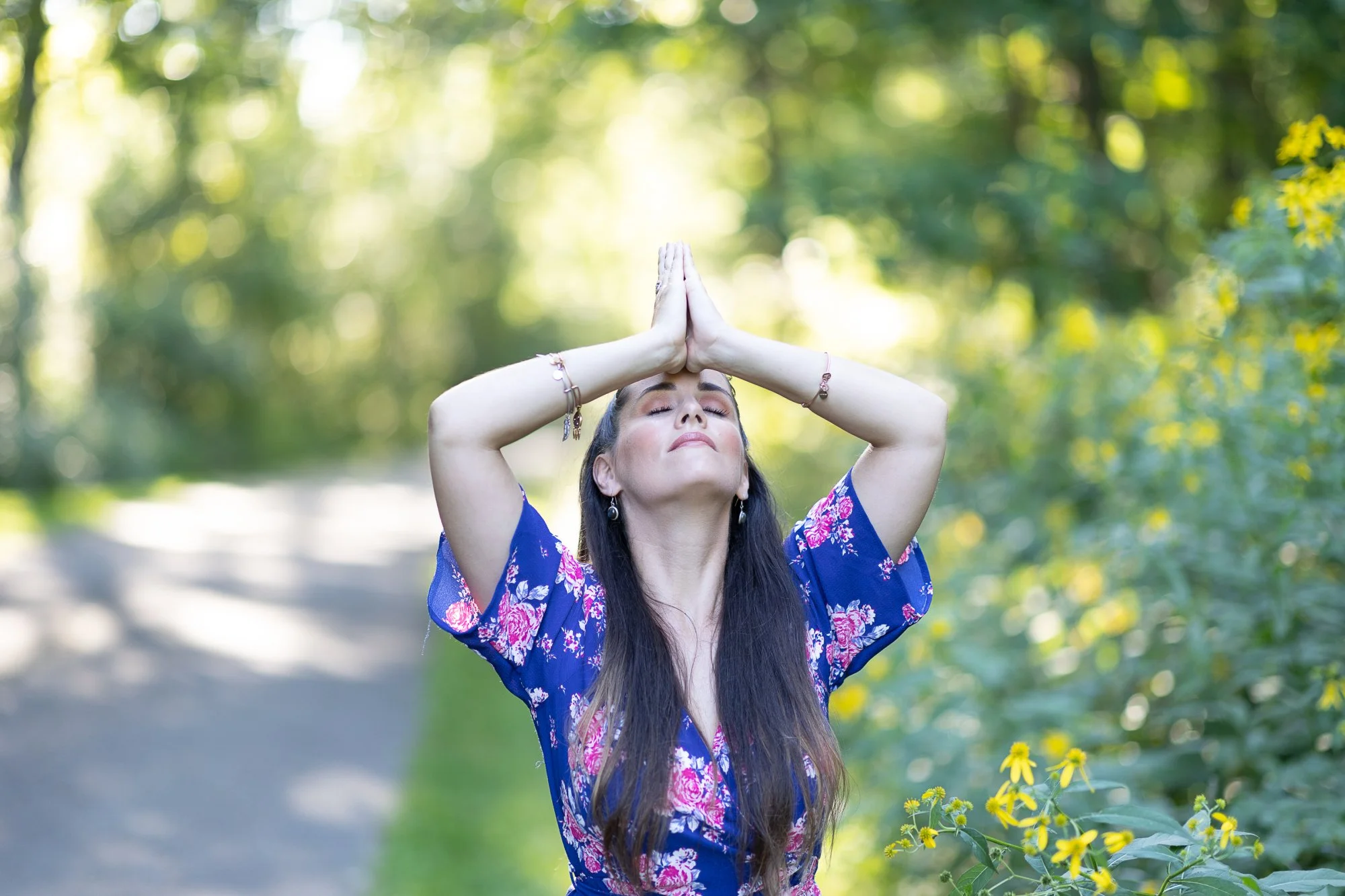 A woman, Alicia, with long dark hair and wearing a blue floral dress is outdoors with her eyes closed, hands pressed together above her forehead, practicing yoga or meditation amidst greenery and flowers.