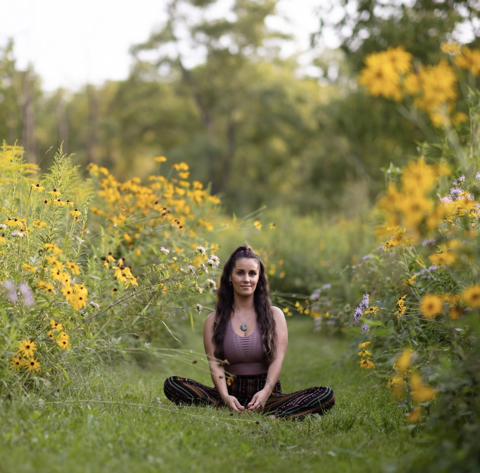 A woman with long dark hair sitting cross-legged in a grassy area surrounded by yellow and some purple flowers, with a blurred background of green trees and bushes, during daytime.