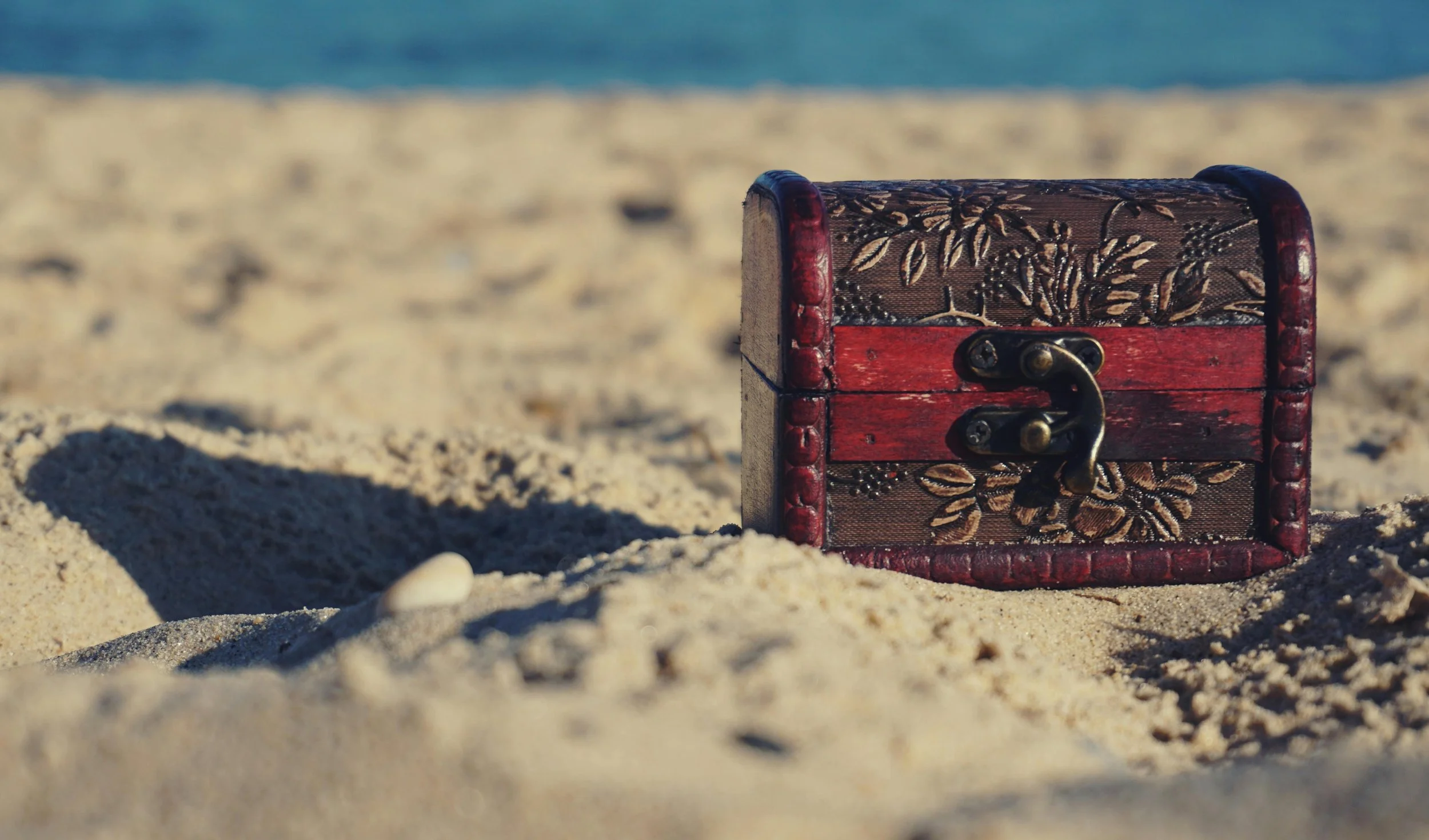 A small decorative wooden treasure chest with floral patterns on the lid, partially buried in sandy beach.