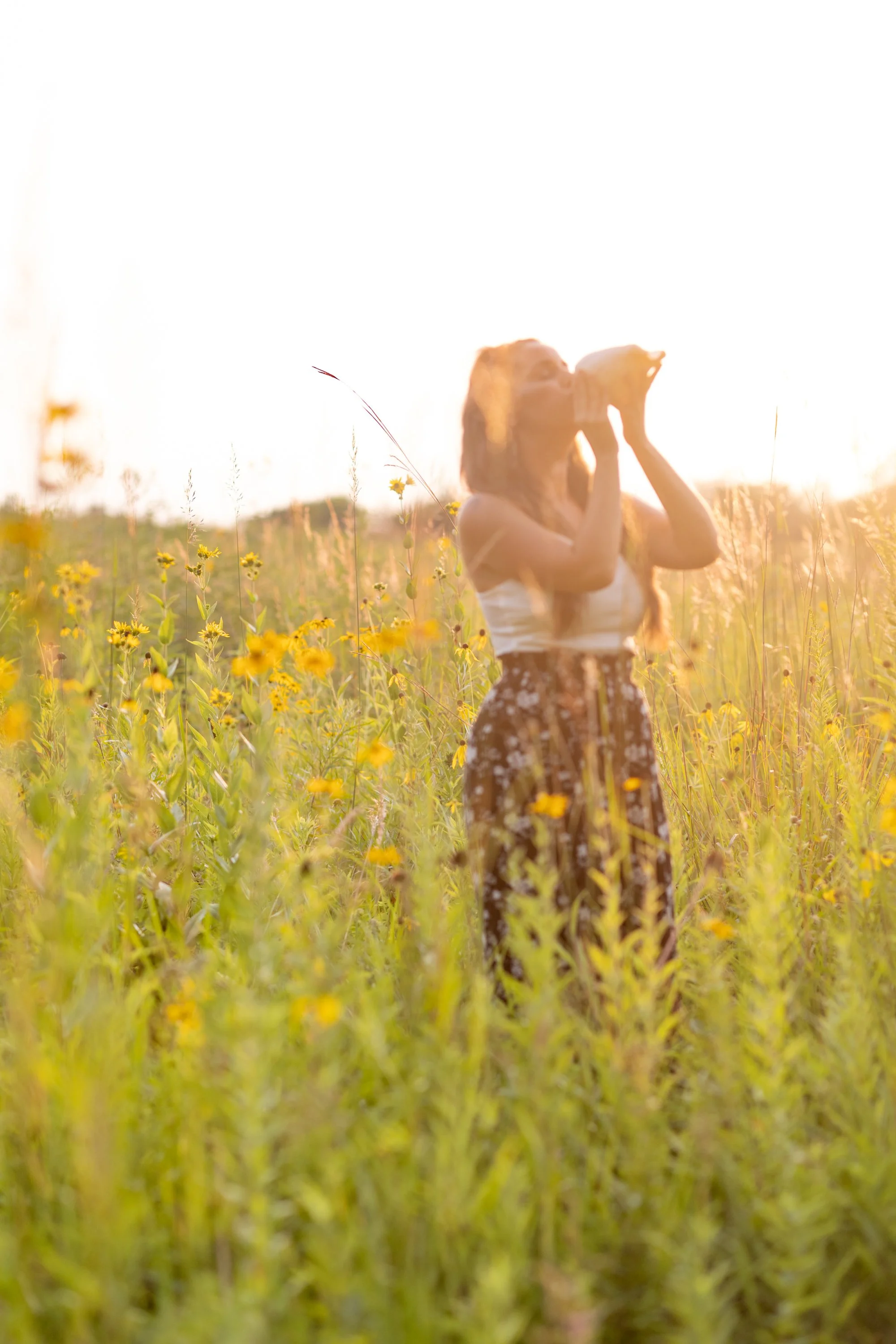 A woman with long blonde hair standing in a field of yellow flowers, drinking from a glass jar during sunset.