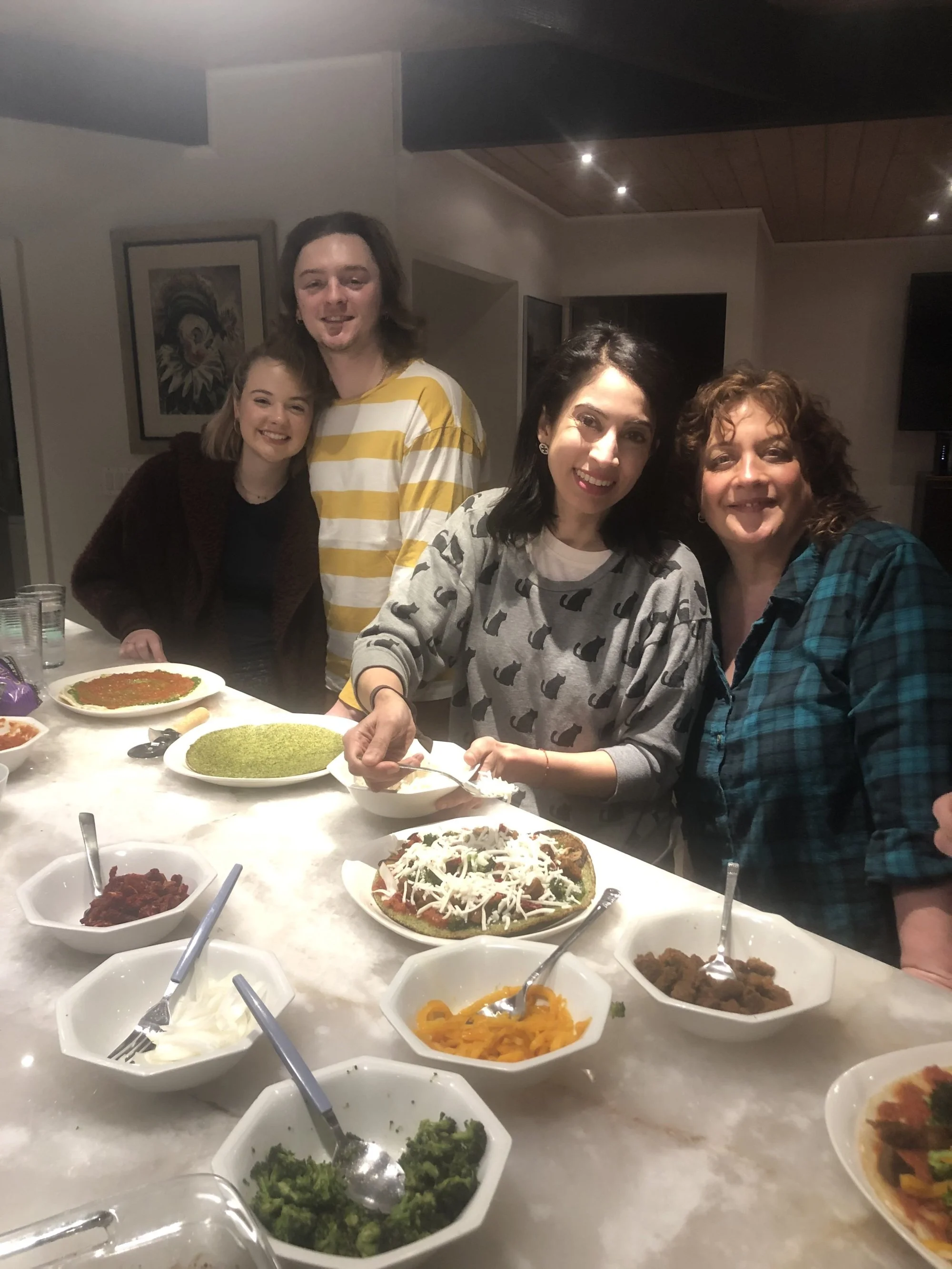 A group of four women smiling and posing behind a table filled with various dishes, including pizzas, salads, and side dishes, in a cozy indoor setting.