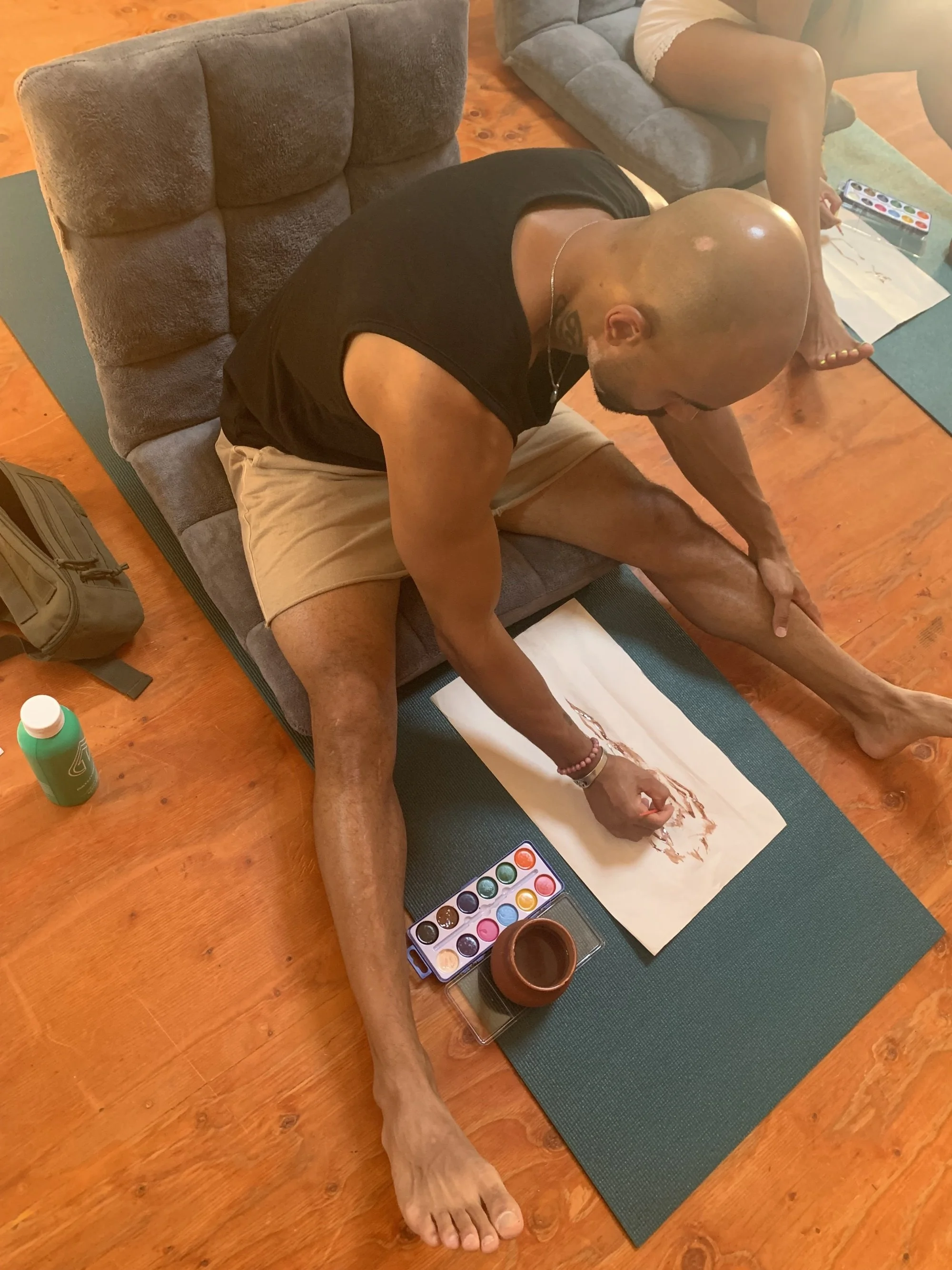 A man with a shaved head practicing yoga on a mat, sitting with one leg extended and the other bent, reaching forward to hold his foot. There are watercolor paints, a paintbrush, a small bowl, and a sheet of paper nearby.