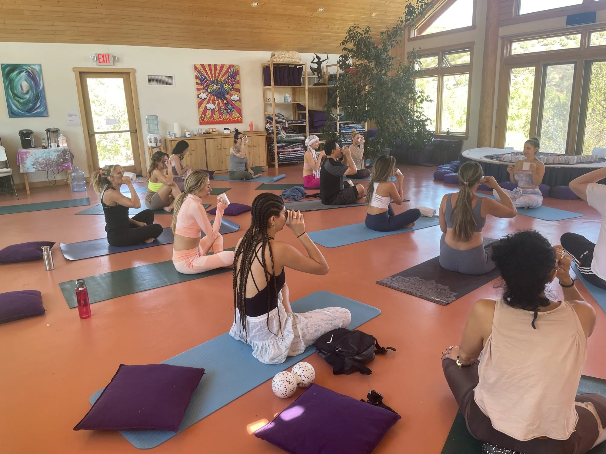 A group of people participating in a meditation or yoga class in a spacious, sunlit room with large windows, artwork on the walls, and mats on the floor.