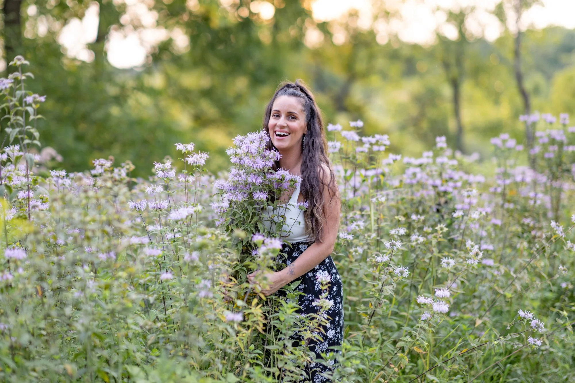 A woman with long wavy hair smiling in a field of purple flowers with a lush green forest background, during daytime.