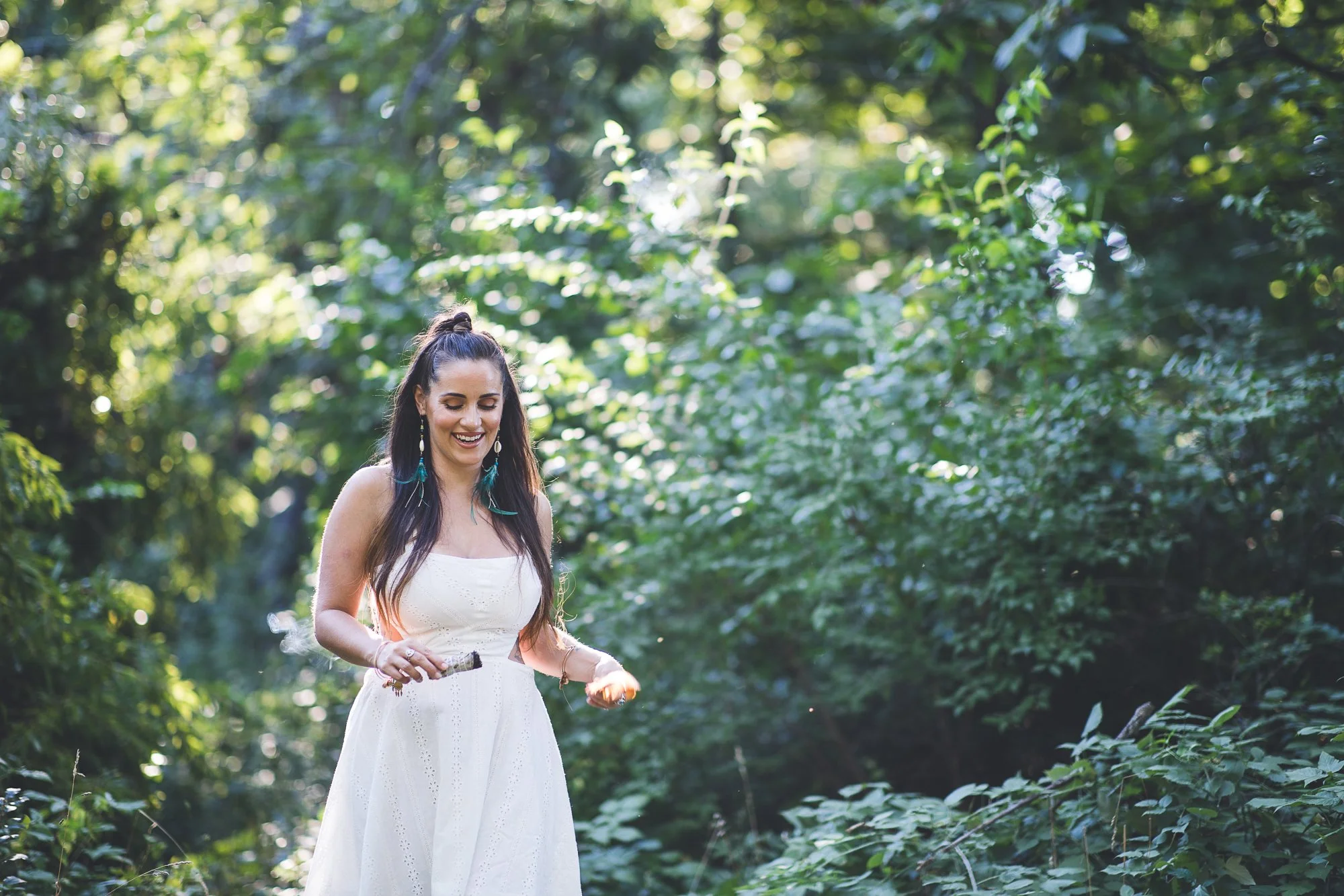 A woman, Alicia, in a white dress standing in a lush green forest, smiling and looking down.