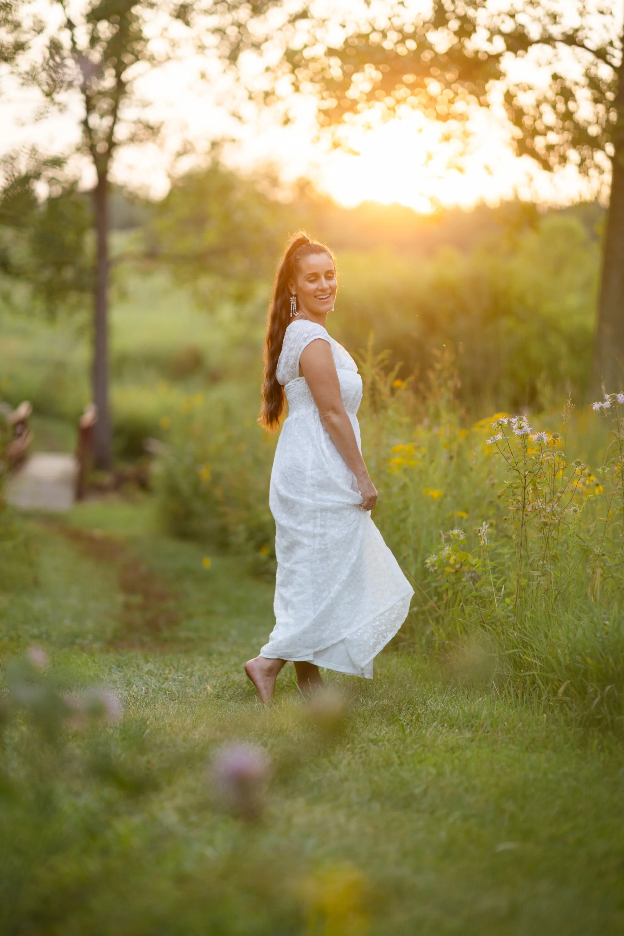 A smiling woman, Alicia in a white dress walking barefoot on grass in a garden during sunset.