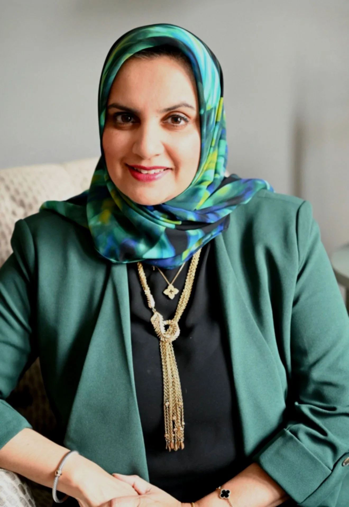 A woman wearing a colorful headscarf, green blazer, and jewelry, sitting indoors and smiling at the camera.