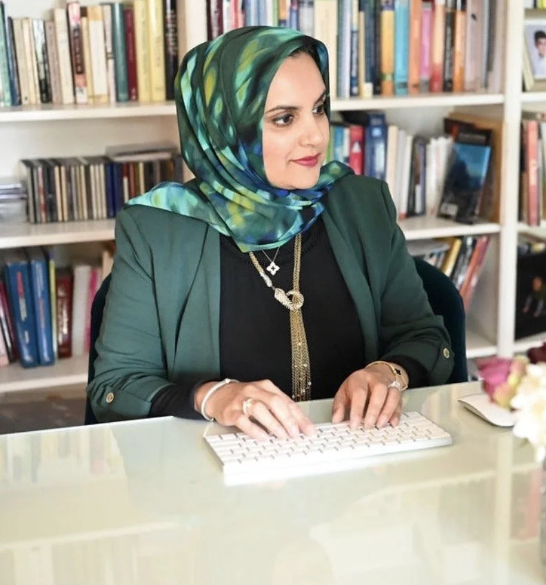 A woman wearing a multicolored headscarf and a green blazer working at a computer in a library or office setting.
