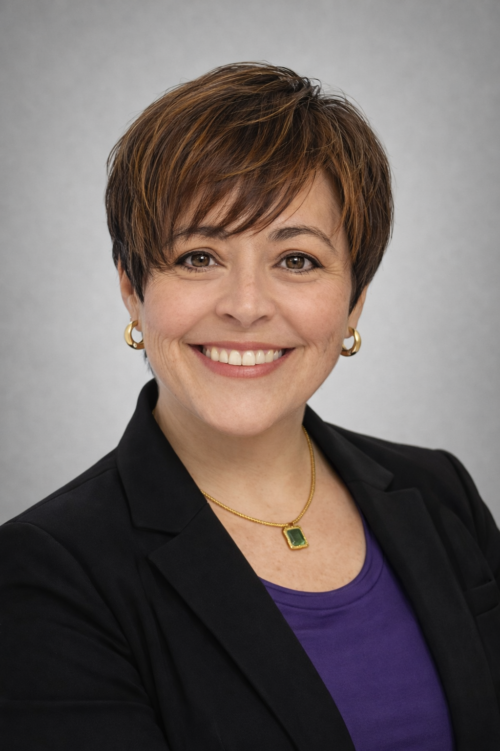 Smiling woman with short brown hair wearing earrings, a necklace with a green gemstone, a purple top, and a black blazer.