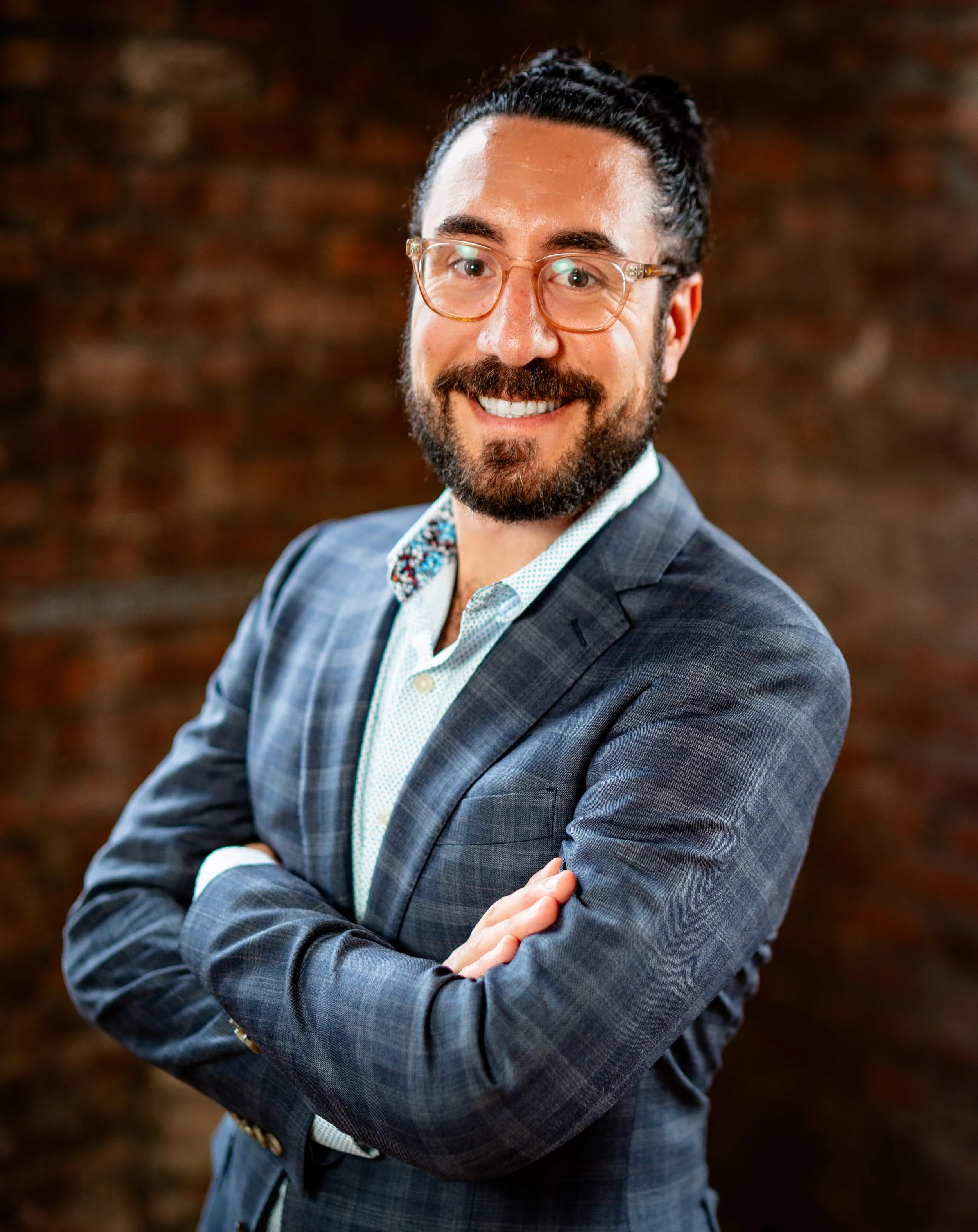 A smiling man in a plaid suit with glasses, crossed arms, and a light-colored shirt, standing in front of a blurred brick wall background.