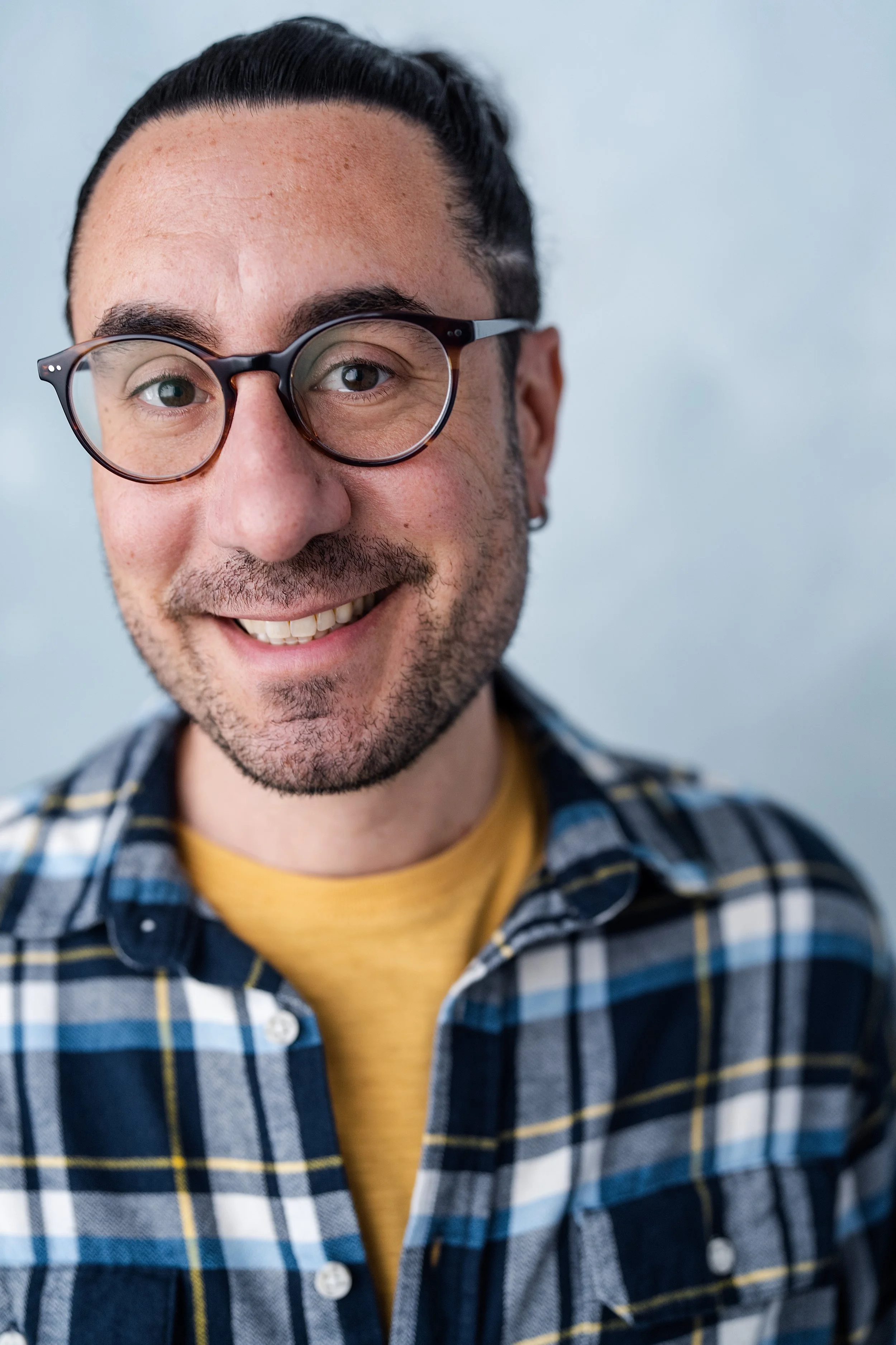 Close-up of a smiling man with glasses, dark hair pulled back, light skin with freckles, and wearing a blue and white plaid shirt over a yellow t-shirt, against a plain light blue background.