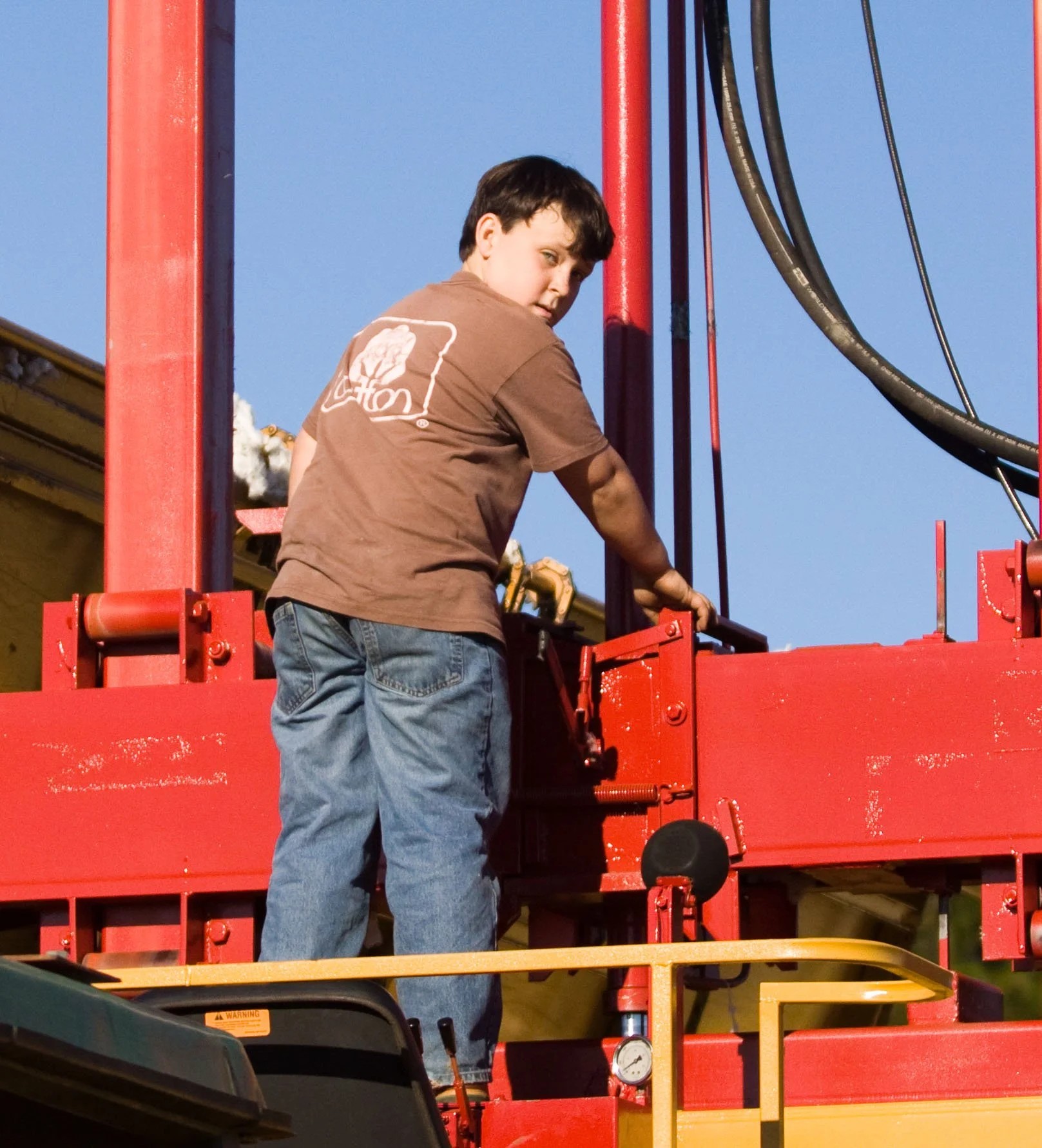 A young boy with dark hair wearing a brown T-shirt and blue jeans is on a red tractor black cables attached to the structure, against a clear blue sky.