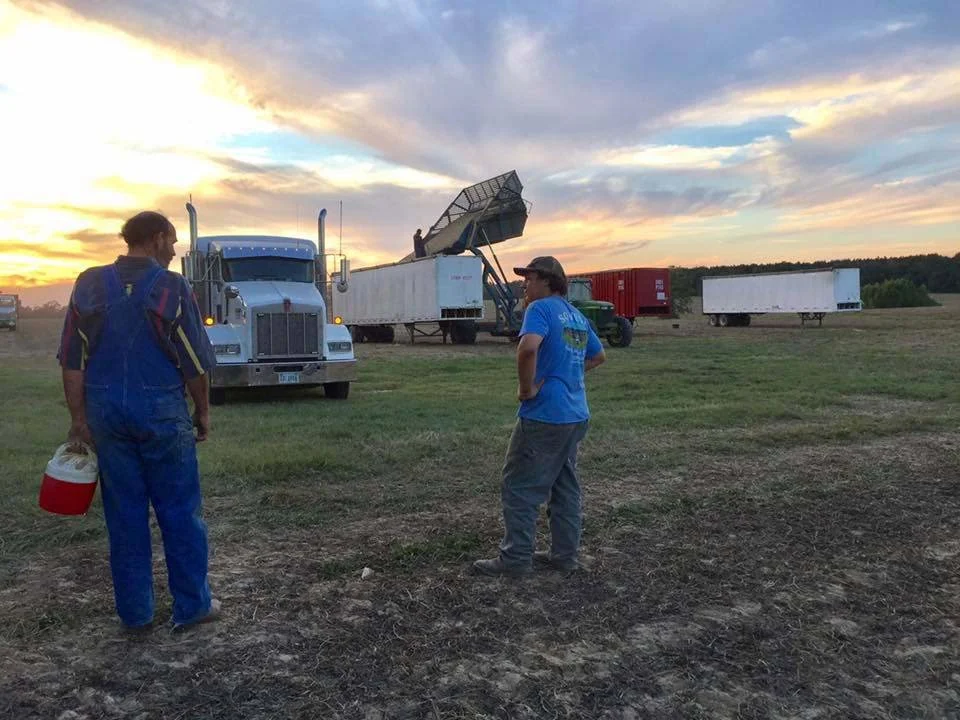 Two men standing in a grassy field at sunset, near trucks and farm equipment, with a partly cloudy sky.