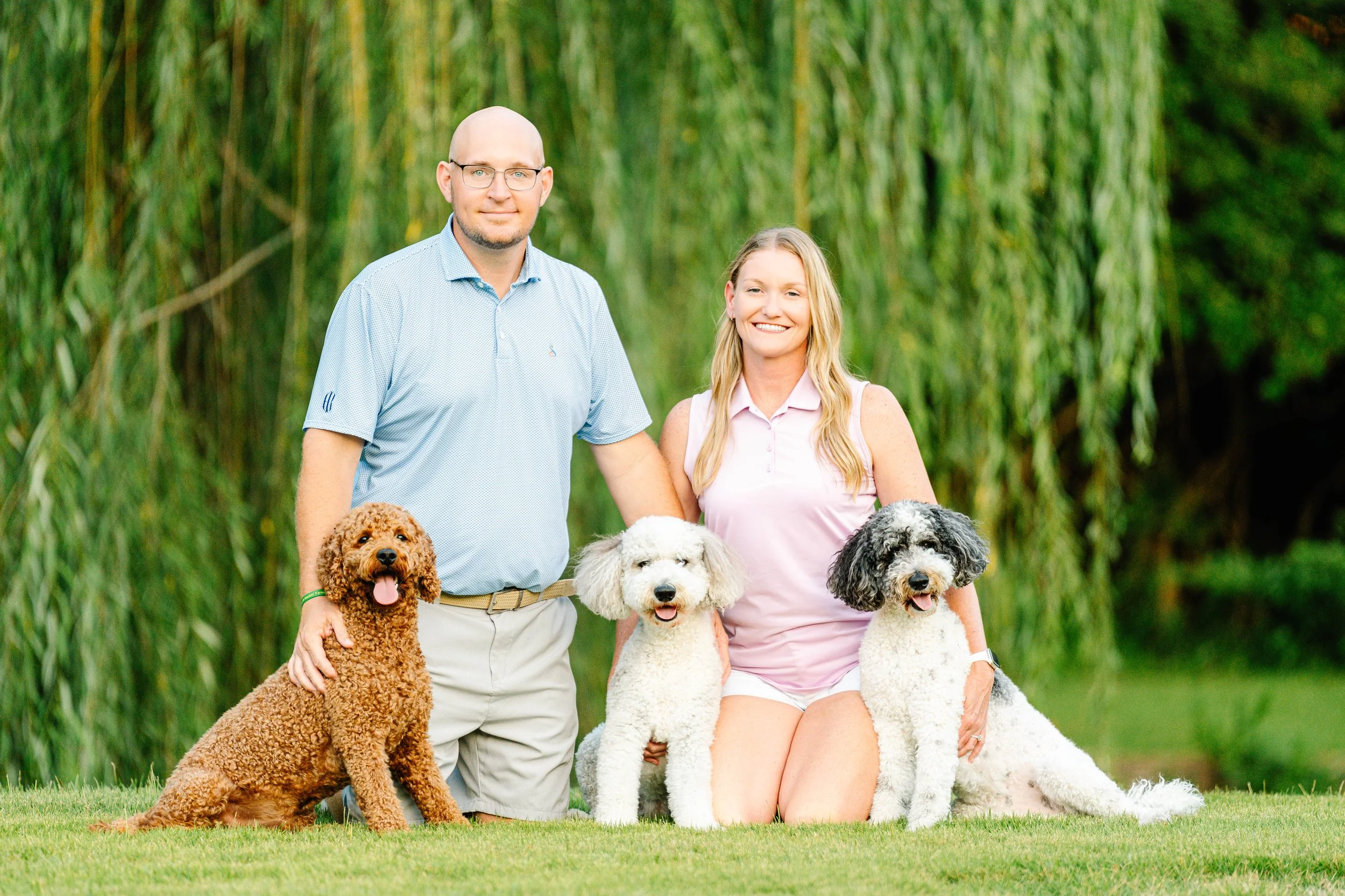 Crystal Holt and husband are outdoors with five dogs, sitting and standing on grass with trees in the background.