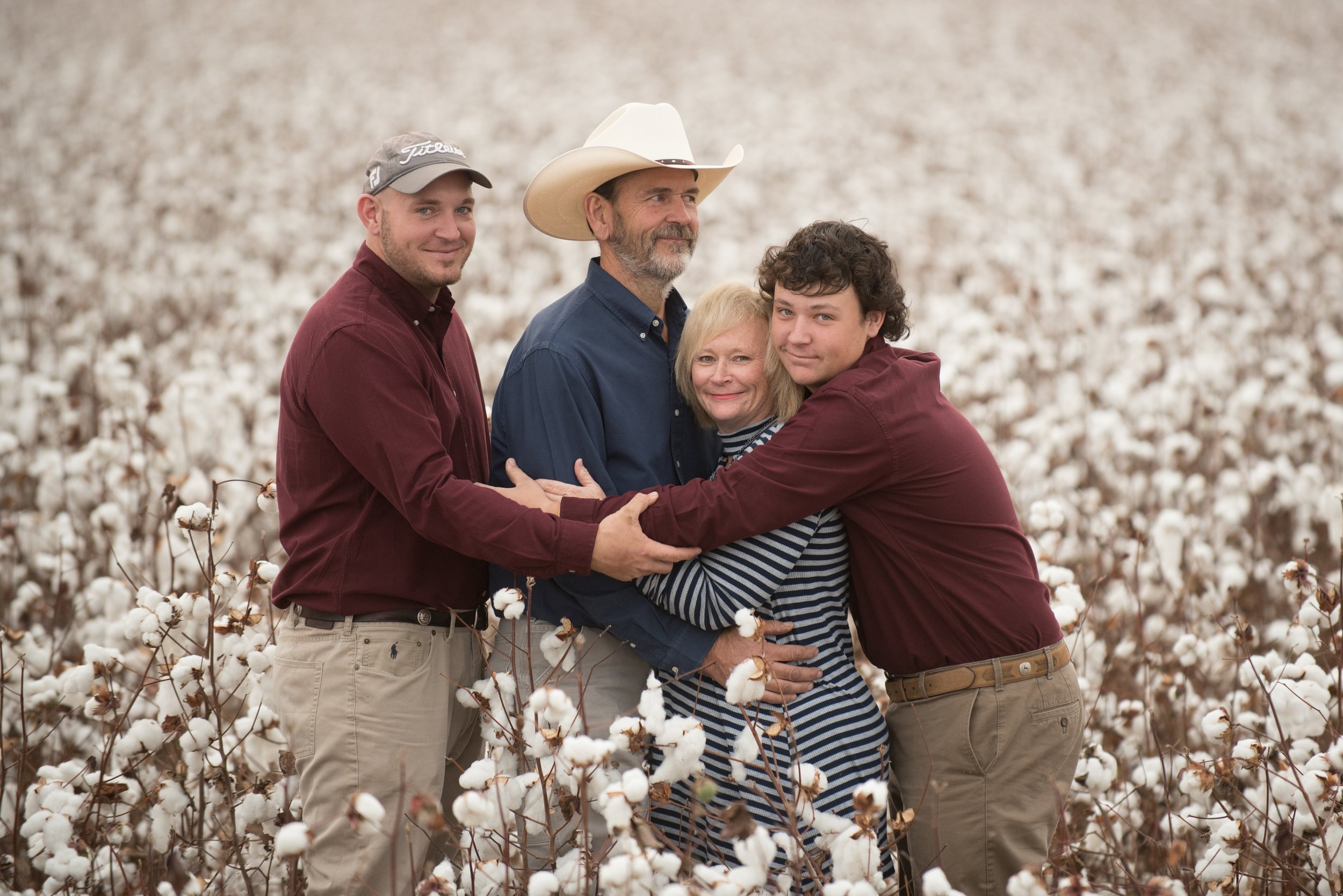 Tammy Holt and her family embracing in a cotton field, smiling at the camera. They include three men, one woman, and one younger male, with a blurred background of white cotton plants.