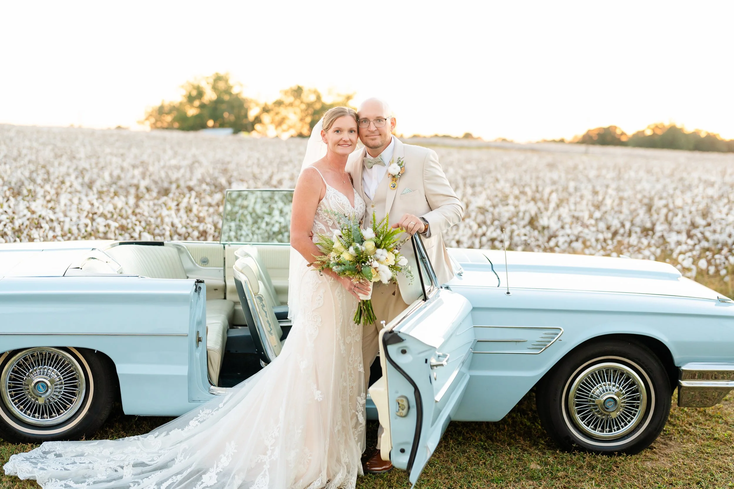 A bride and groom standing beside a vintage light blue convertible car in a field of cotton, during sunset. The bride is holding a bouquet and wearing a lace wedding dress, while the groom is dressed in a beige suit and bow tie.