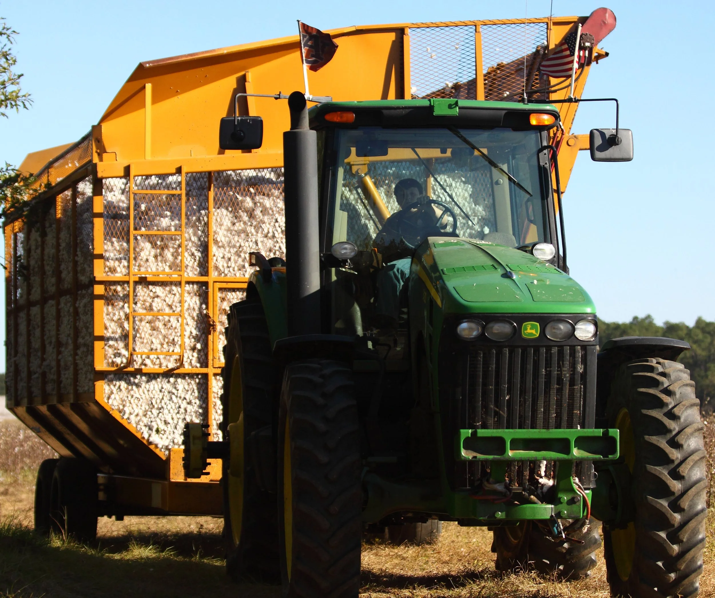 A green John Deere tractor with an orange harvesting attachment on a farm field, under a clear blue sky.