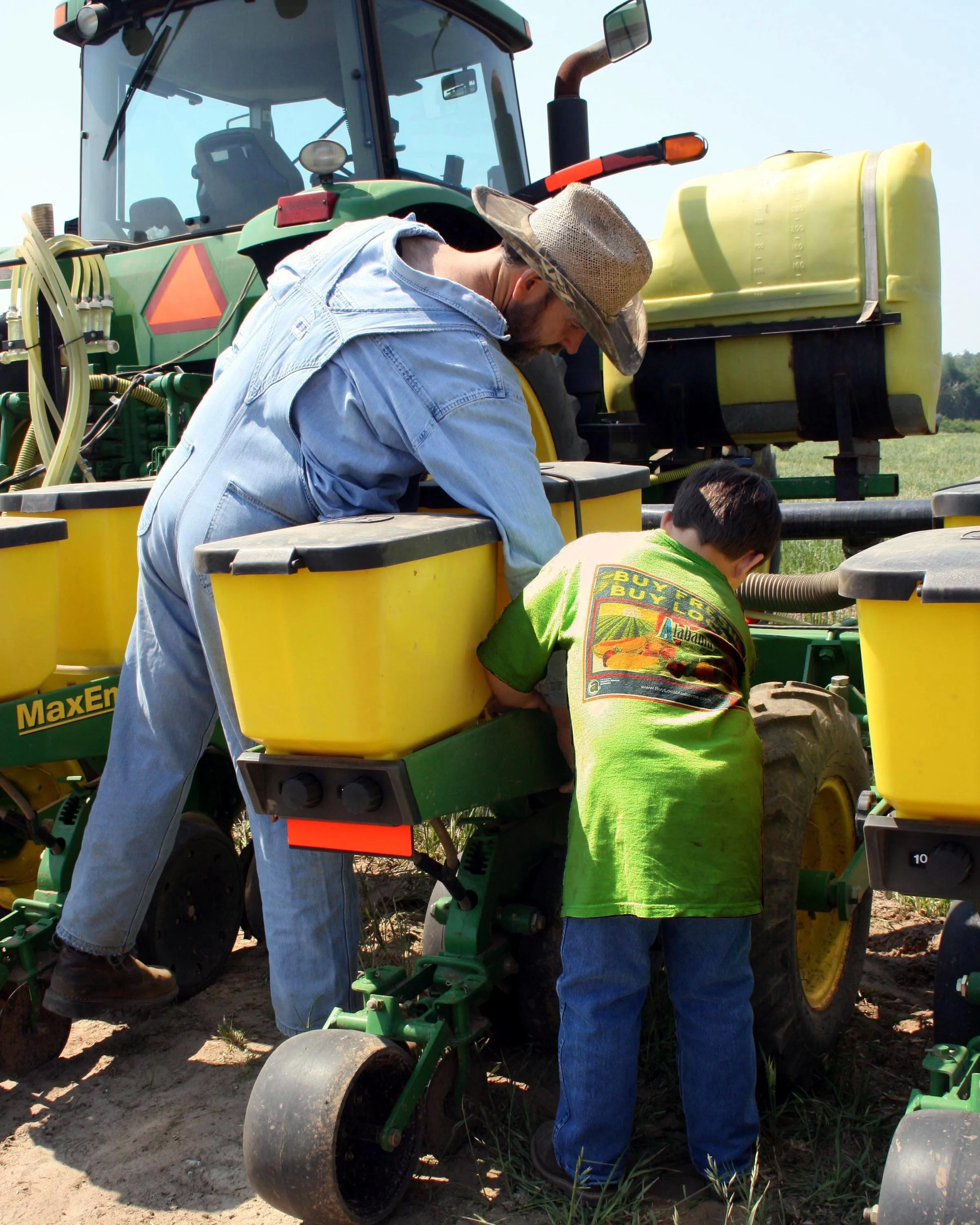 A man wearing a straw hat, blue denim shirt, and blue jeans is working on a green and yellow tractor in a field. Two children, one in green and the other partially visible, are nearby. The man is leaning into the machinery, possibly repairing or inspecting it. The tractor features a large yellow tank and various mechanical parts.