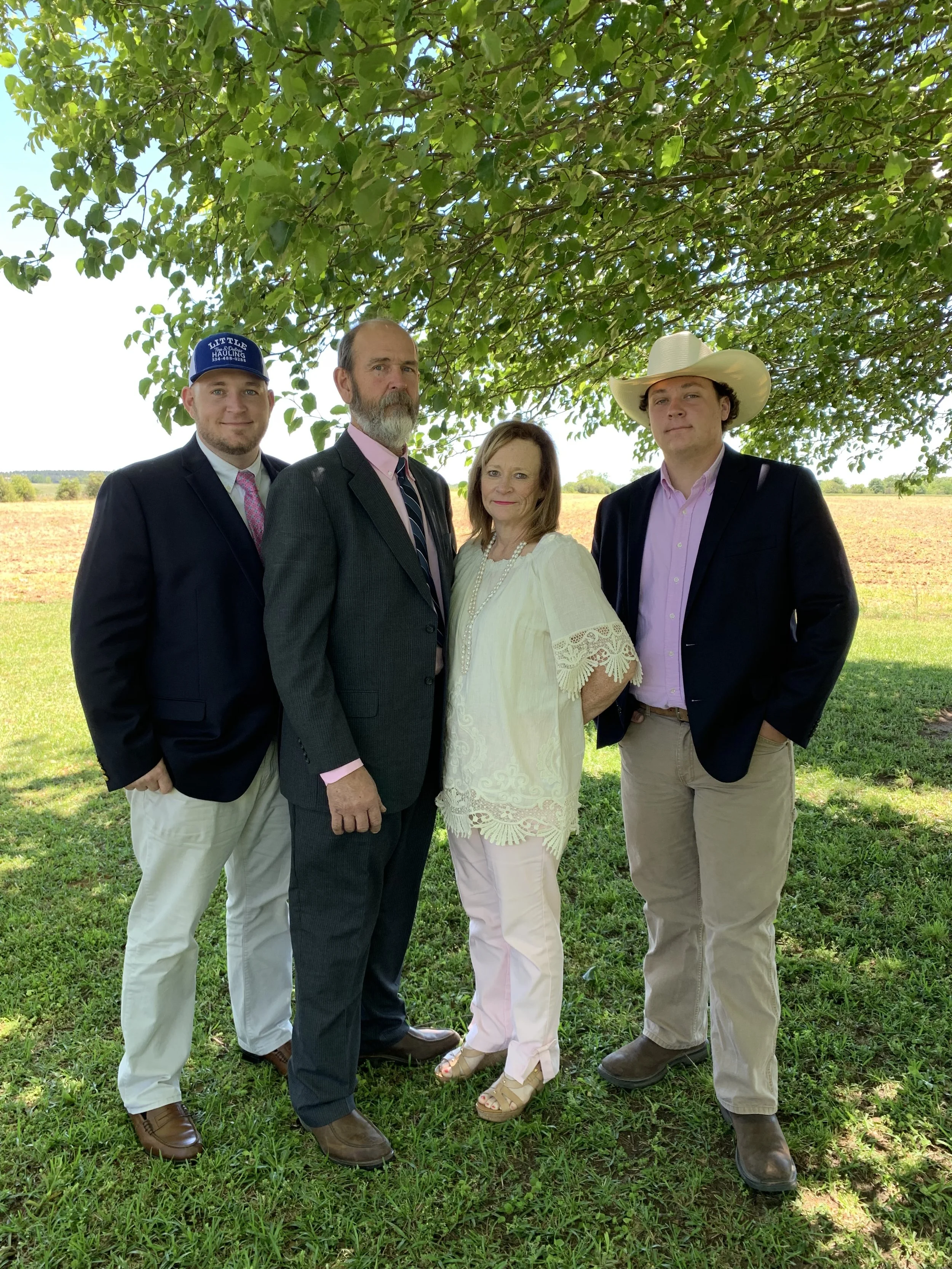 The Holt family of four standing outdoors under a large leafy tree on a sunny day, with a field in the background. They are dressed in semi-formal attire, with two men in suits, a woman in a white blouse, and a young man wearing a cowboy hat.