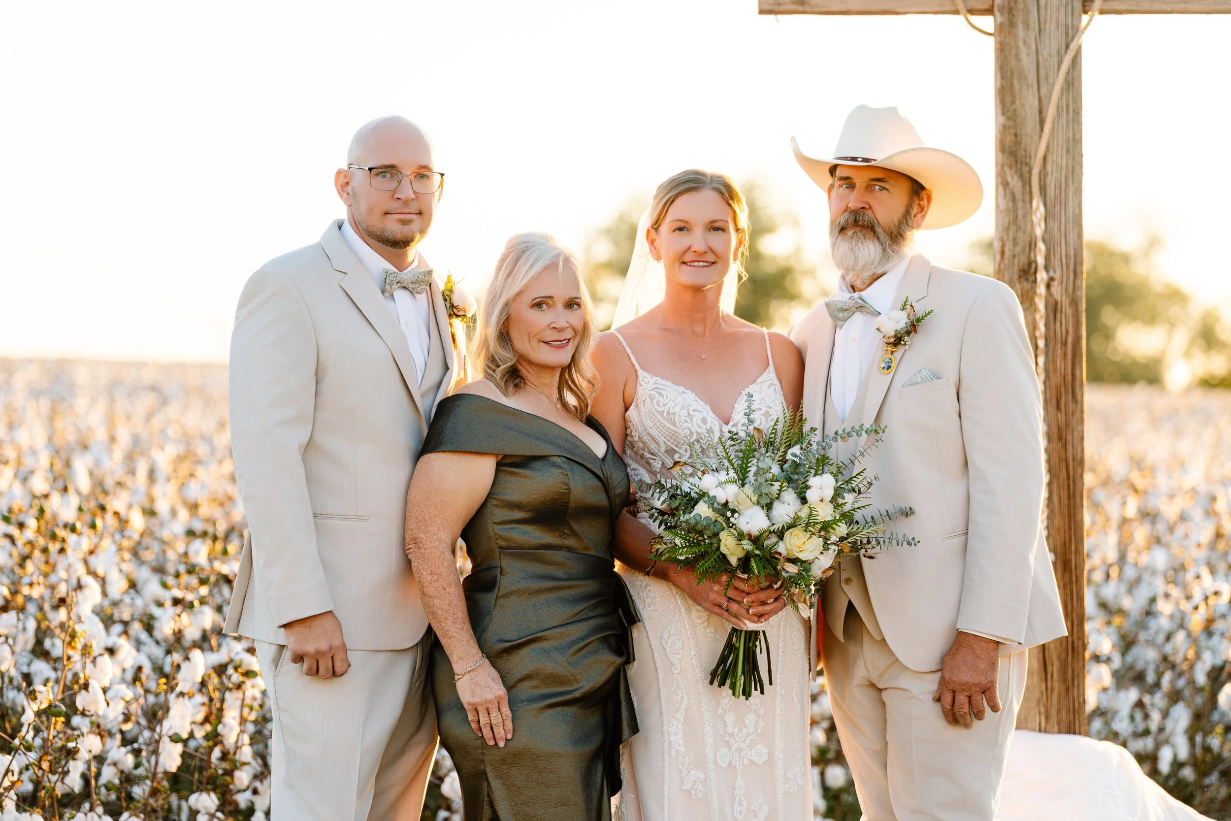 A group of five people at a wedding photo, including a bride in a lace wedding dress holding a bouquet, two men in light-colored suits with boutonnières, and two women, one in a dark dress and one in a black dress, standing outdoors near a wooden pos