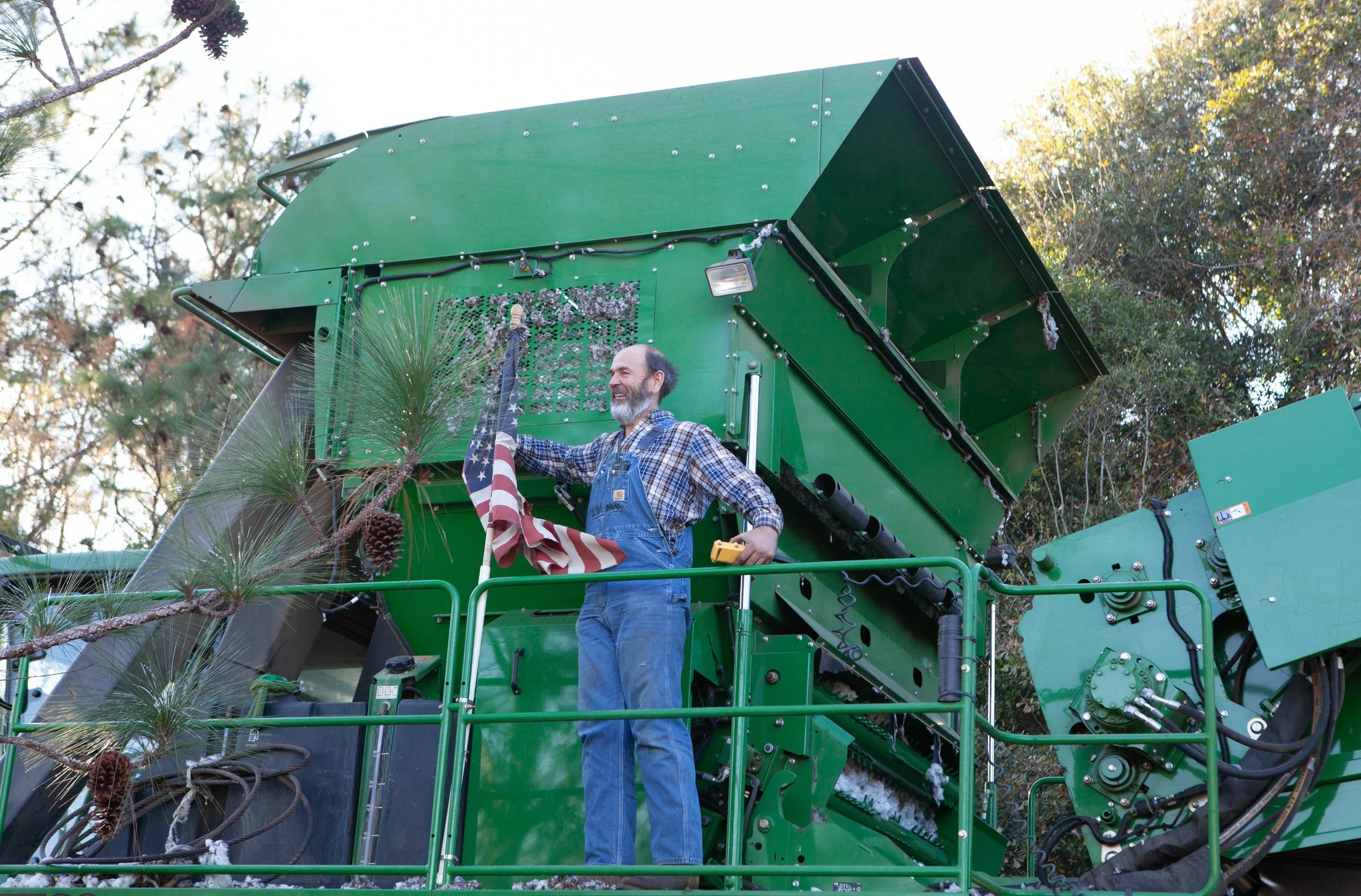 A man with a beard and glasses in blue overalls standing on a green machine outdoors, holding a flag with stars and stripes, with trees and sunlight in the background.