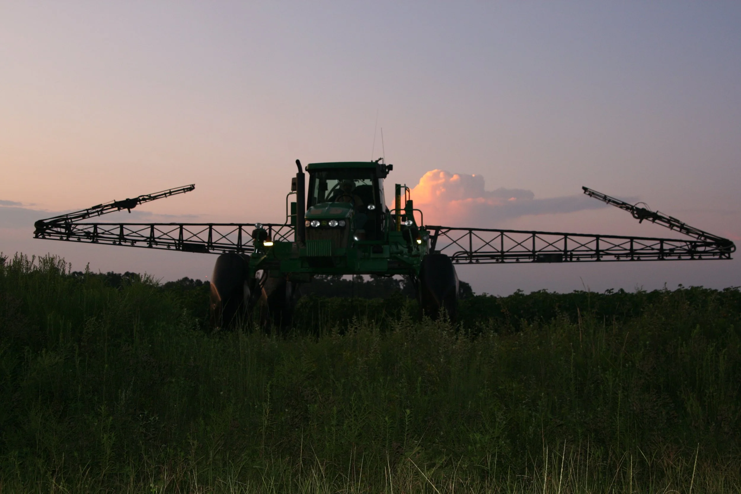Green tractor with a wide spray boom attachment operating in a field at sunset, with a partly cloudy sky in the background.