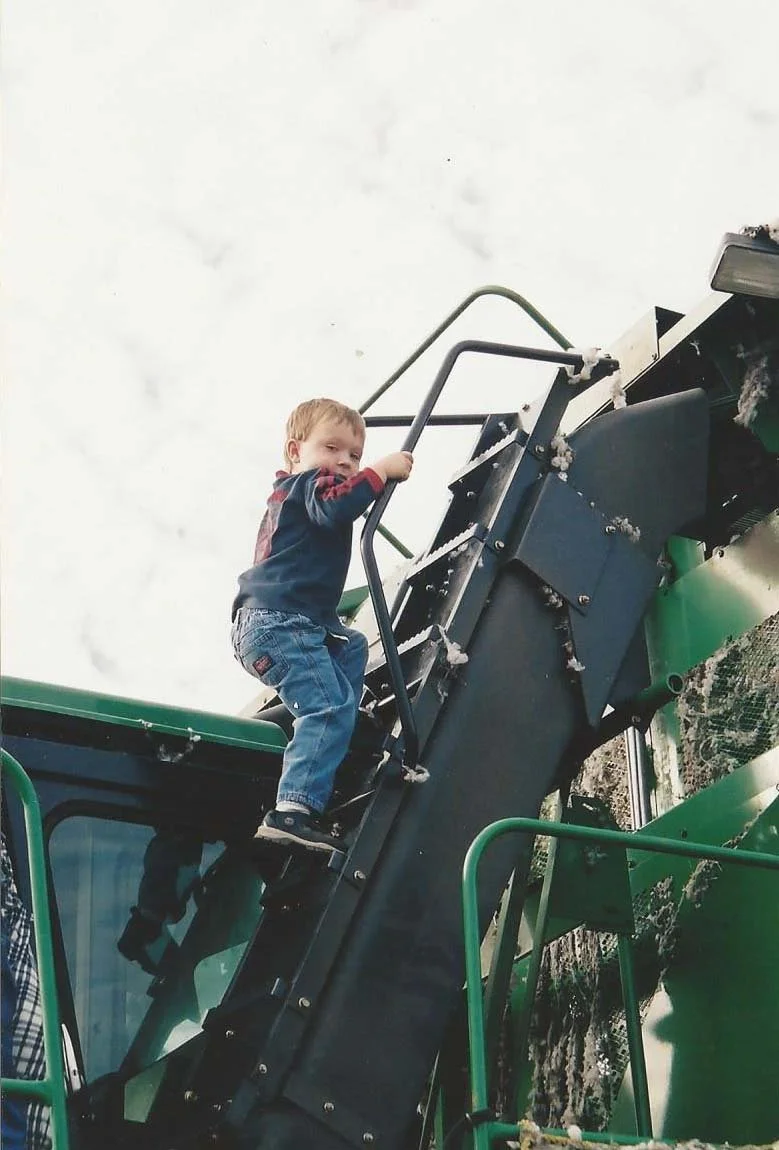 A young boy is climbing up a ladder attached to a large green and black outdoor cotton gin, with cotton and cloudy sky in the background.