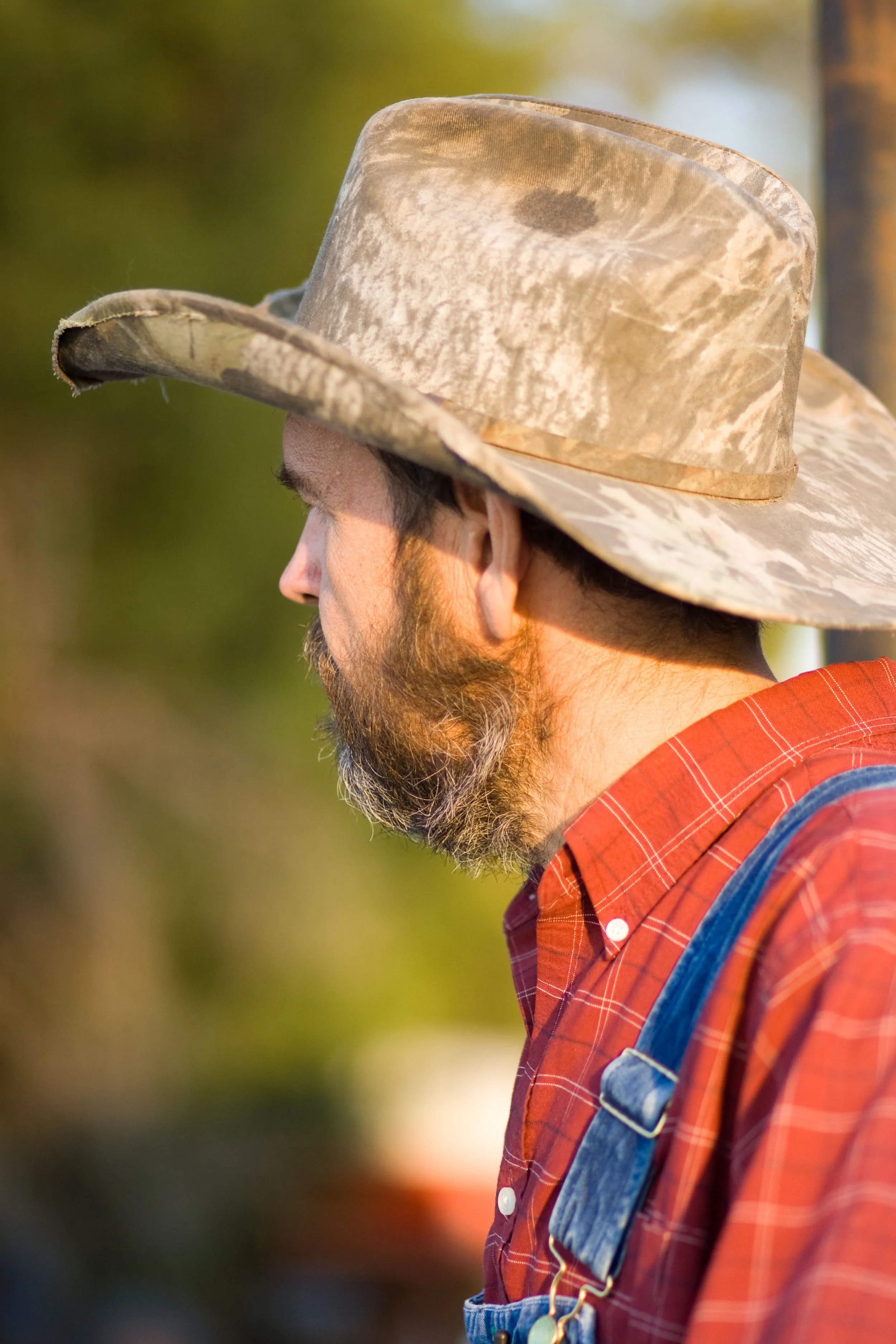 Close-up photo of a man wearing a camouflage hat, a red plaid shirt, and a denim overall, with trees and blurred background.