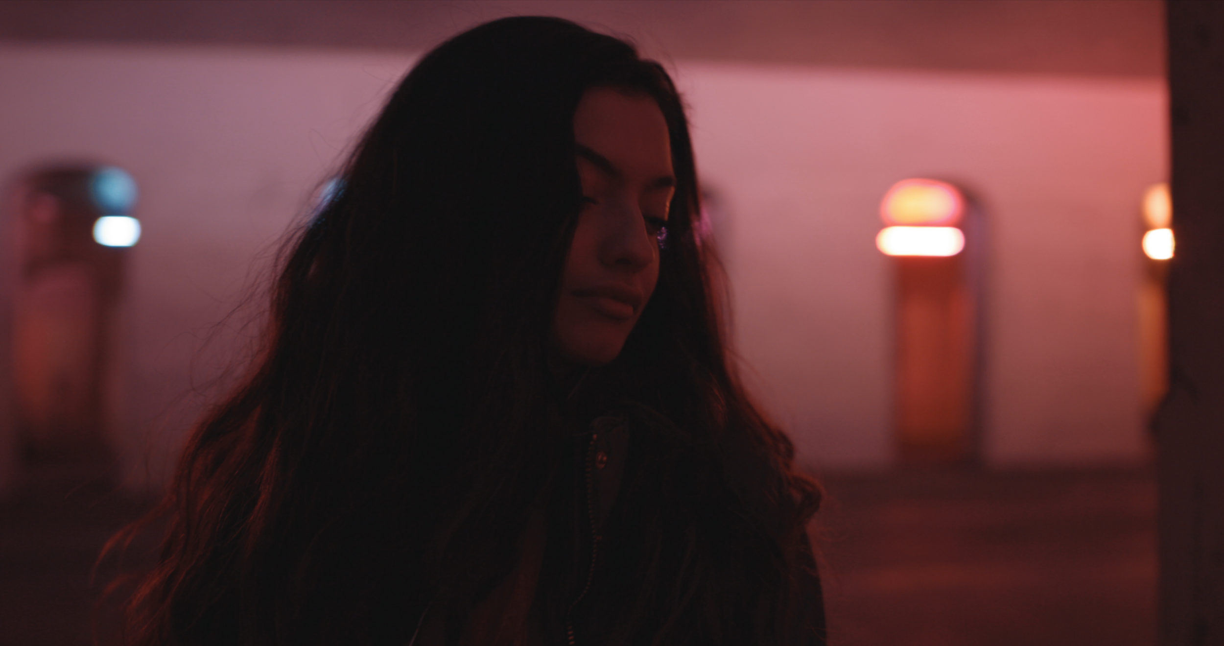 Silhouetted person with long hair in dimly lit room with pink, purple, and red lights.