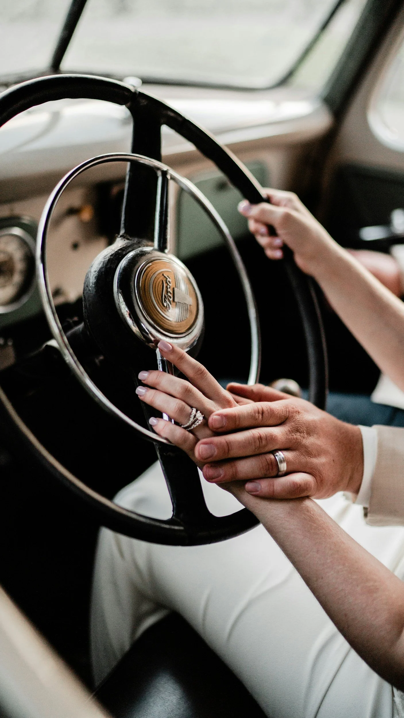 A person with wedding rings on their fingers holding hands with another person and resting on the steering wheel of a vintage Ford vehicle.