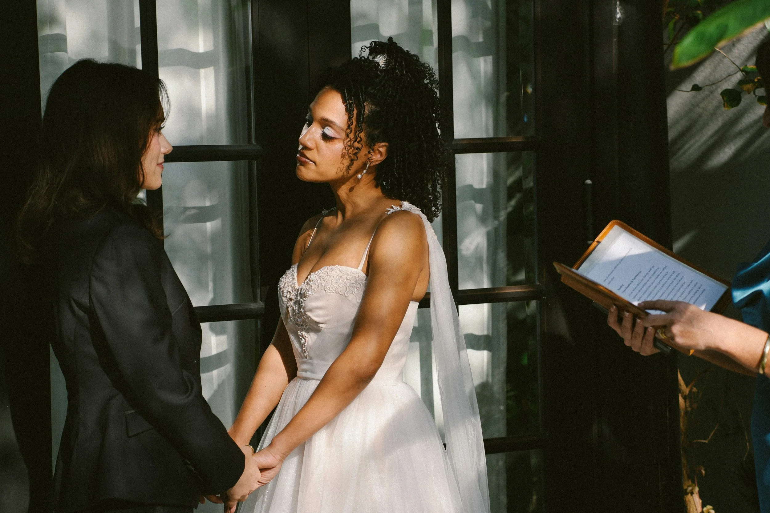 A wedding ceremony with two women holding hands and facing each other, one wearing a white wedding dress and the other in a dark suit, while an officiant holds a book and prepares to conduct the ceremony.