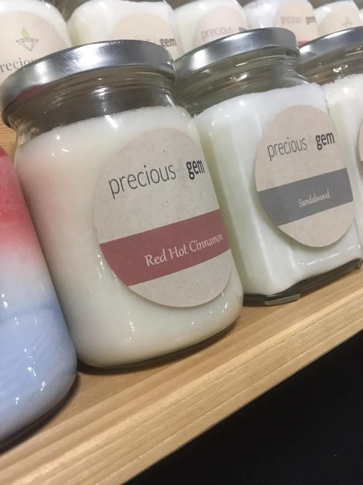 A glass jar candle labeled 'precious gem' with a scent of Red Hot Cinnamon, placed on a wooden shelf.