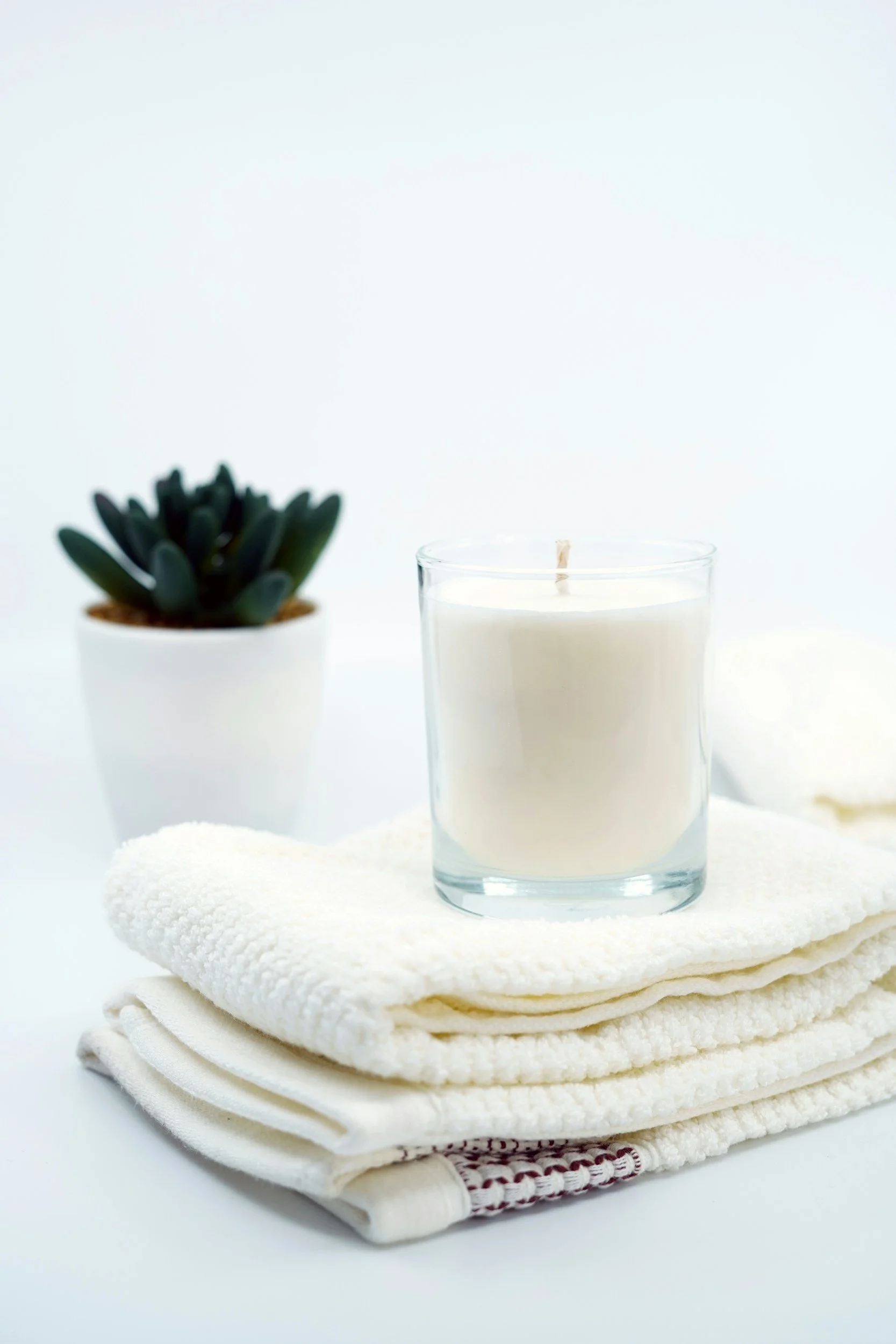 A clear glass candle with a wick, placed on a folded cream-colored towel, with a small potted succulent plant nearby, all on a white background.