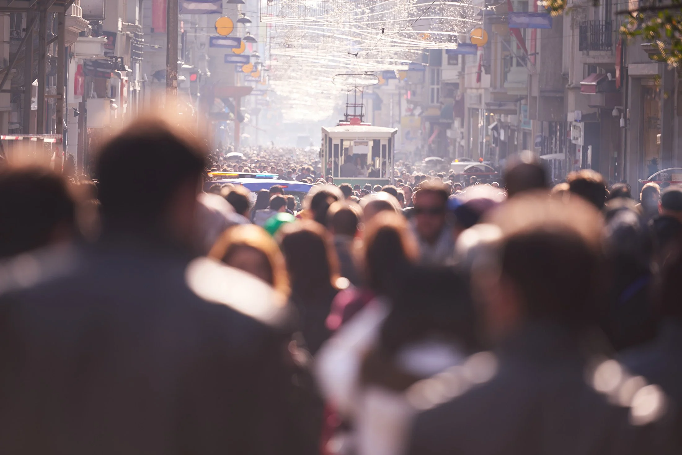 A crowded urban street scene with many people, a trolley, buildings, and overhead wires, during daytime.