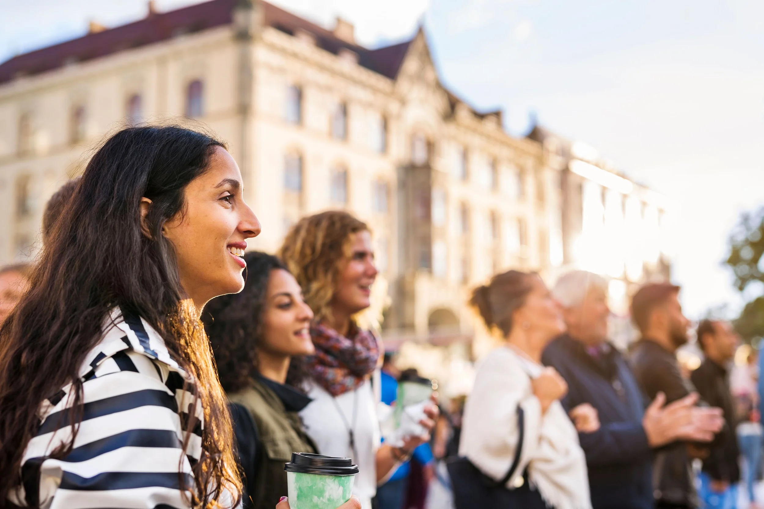 A group of people standing outdoors in front of historic buildings, with a woman in the foreground holding a green coffee cup, smiling and looking to the left.